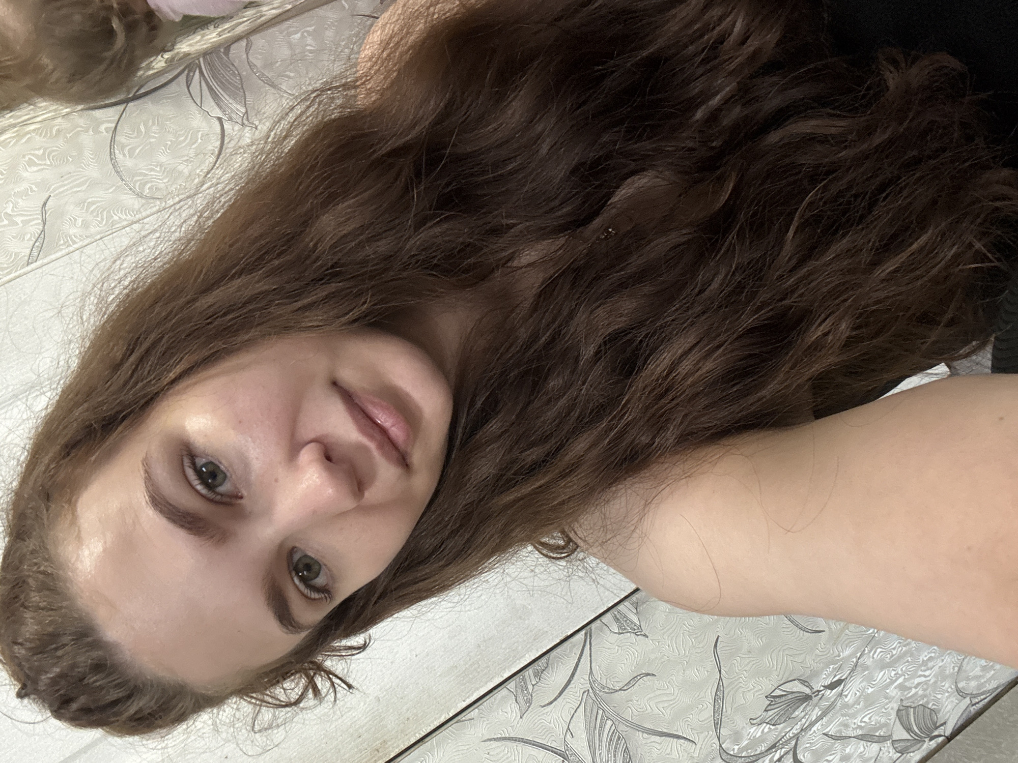 A woman with long, wavy brown hair is lying on a surface with a floral pattern. Her face is visible, showing light skin, green eyes, and natural makeup. Part of her arm is visible in the lower right corner of the image. The image appears to be a selfie taken from above. There is no indication of a location in the city of Nizhnyaya Gar', Russia in this image.