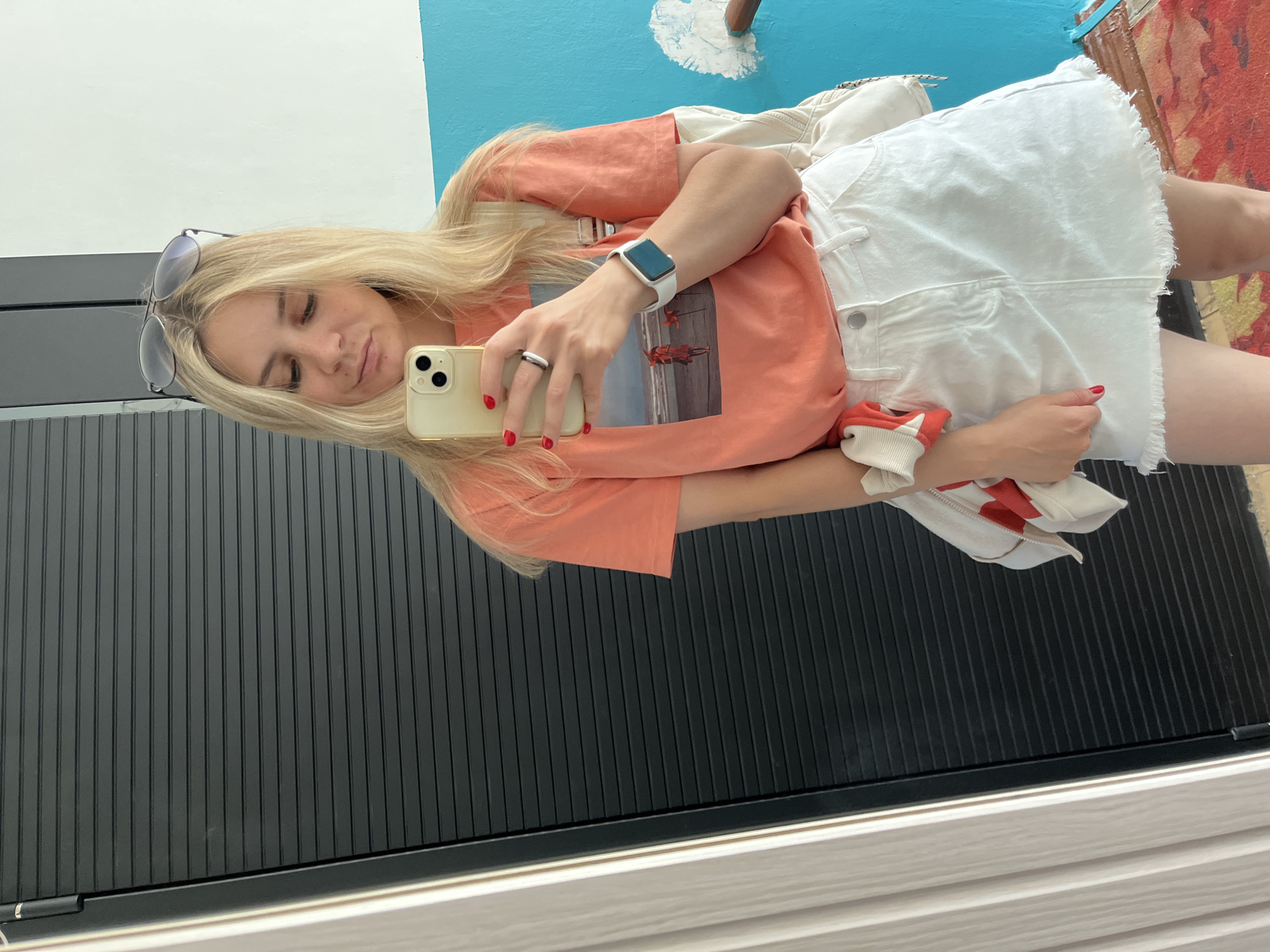 A young blonde woman with sunglasses on her head takes a selfie in front of a black ribbed door and a blue wall. She is wearing an orange t-shirt with a beach scene graphic, a white denim skirt, and a white backpack. She is also wearing a white smartwatch on her left wrist and a silver ring on her right index finger. A red and white striped jacket is draped over her left arm. The floor is covered with a patterned rug. The scene appears to be an interior setting, possibly a hallway or doorway in a residential or public building in Chaikovsky, Russia.