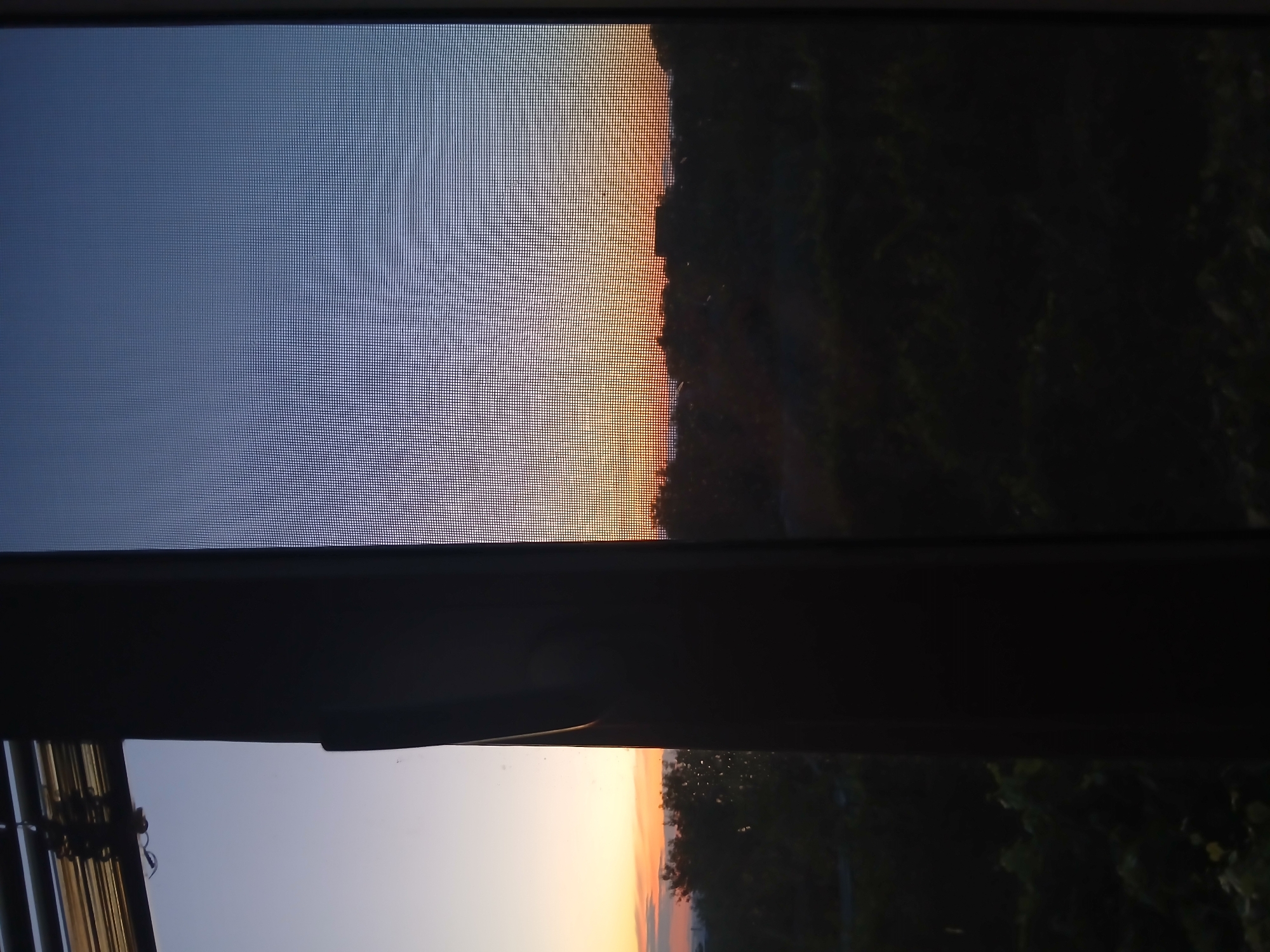 The image shows a view through a window with a screen at sunset. The sky is a gradient of blue and orange. The silhouette of trees and buildings is visible against the sky. The image is taken from a residential building in Dnipro, Ukraine, as suggested by the architecture and landscape.