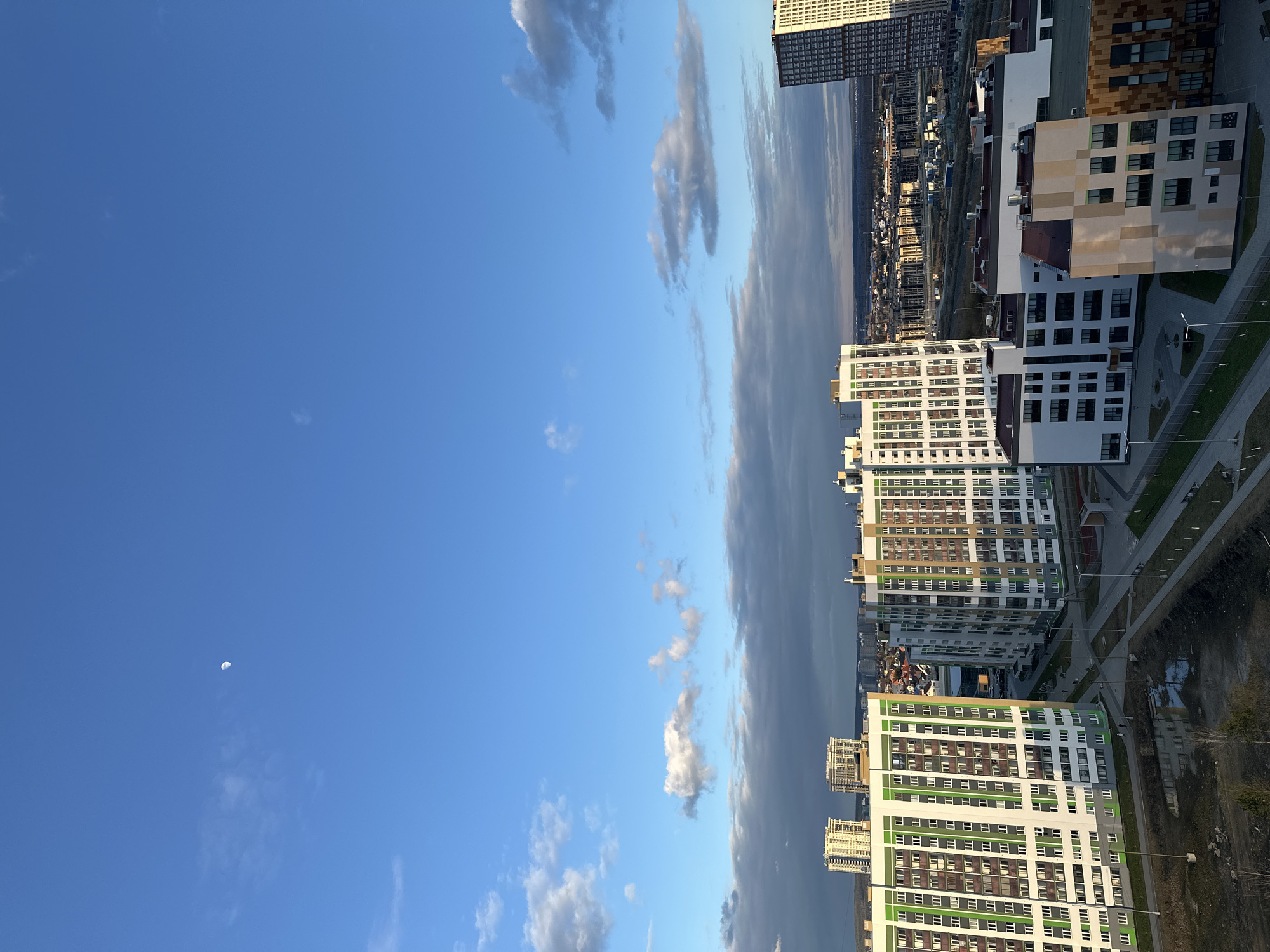 The image shows a view of Yekaterinburg, Russia, with modern apartment buildings of varying colors and heights dominating the lower portion of the frame. The buildings are oriented vertically, with rectangular facades and numerous windows. In the distance, a cityscape stretches out, including more buildings and a glimpse of what appears to be a highway or railway. A clear blue sky covers the upper portion of the image, dotted with light, fluffy clouds. A thin crescent moon is visible in the lower left corner. A large, vertical formation of dark clouds appears to the right of the blue sky. The overall impression is a blend of urban development and natural elements.