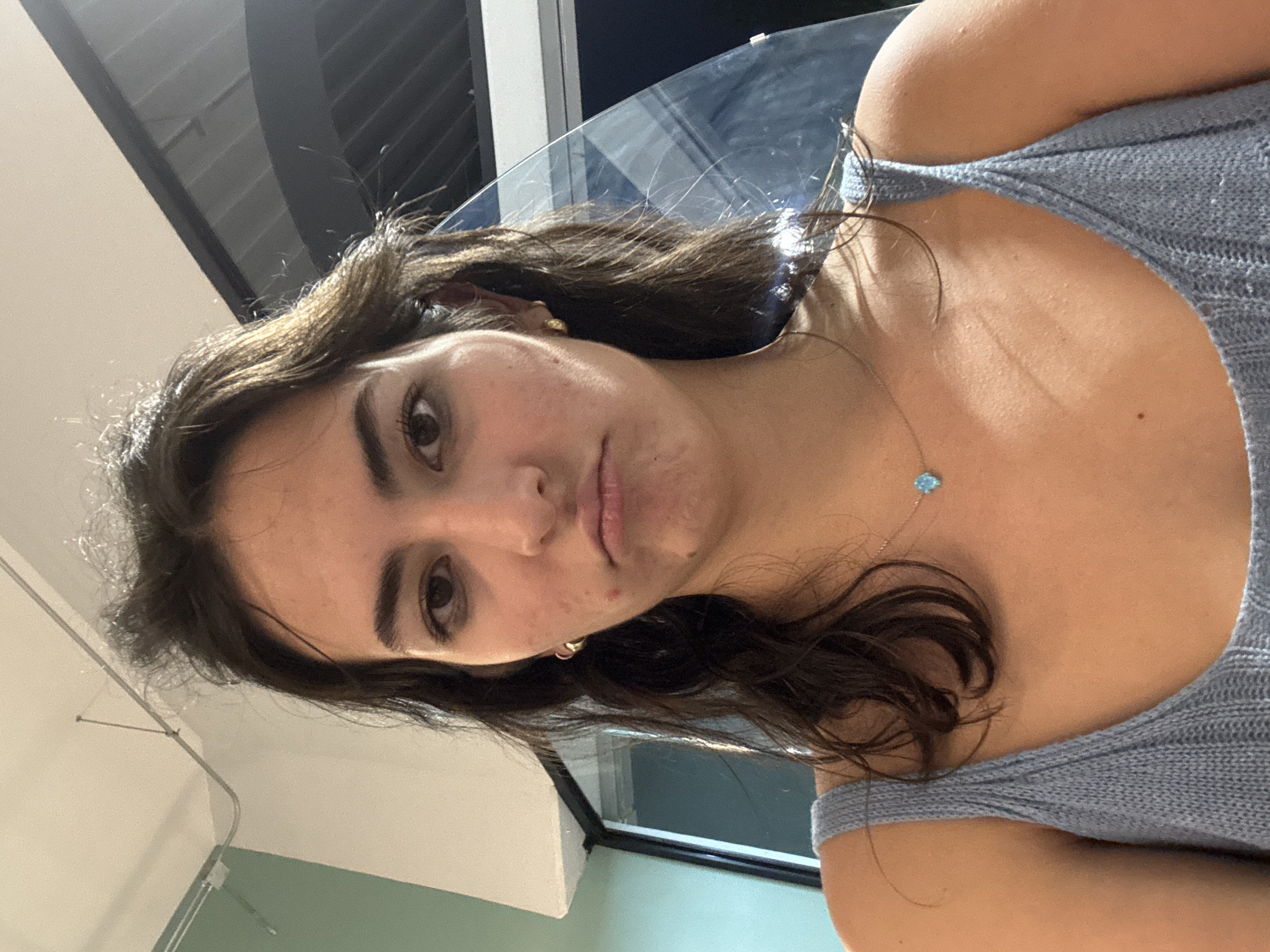 A close-up, rotated portrait image depicts a young woman with a neutral to slightly pensive expression, looking directly at the viewer. Her dark brown, wavy hair frames her face, with strands falling over her shoulders. She wears a light blue or grey knitted tank top, revealing her shoulders and upper chest. A delicate necklace with a small, light blue, possibly hamsa-shaped pendant hangs around her neck, and a small golden hoop earring is visible on her left ear. Minor blemishes are visible on her forehead and chin.
The setting appears to be indoors, with architectural elements forming the background. Behind her, a large, reflective glass surface is prominent, suggesting a window, glass partition, or possibly a table. Further in the background, there are dark rectangular structures that could be windows or vents, and light-colored ceiling or wall panels with a white railing or pipe. The ambient light suggests either an indoor illuminated environment or a nighttime exterior seen through the glass. No specific location cues are visible within the image to confirm it is Puebla, Mexico, other than the provided context. There is no visible text. The overall mood is calm and introspective.
