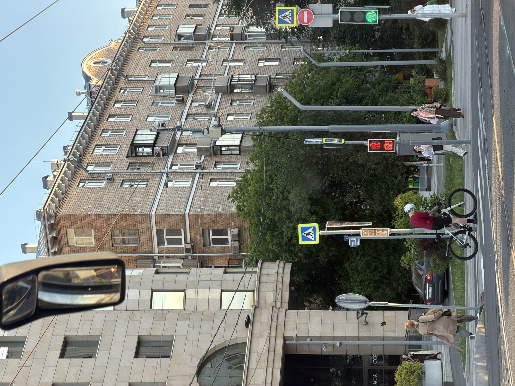This urban scene, set in Moscow, Russia, captures a moment at a bustling street intersection from the perspective of a vehicle. The image is oriented vertically (when corrected from its rotated state), showcasing multi-story buildings, pedestrians, a cyclist, and various street furniture under a clear, bright sky, likely during late afternoon or early evening.
Dominating the background is a large, multi-story brick building with numerous windows, some featuring small balconies or air conditioning units. Its distinctive roofline includes an ornate, rounded dormer window and several vents, suggesting an older, classic architectural style. In the foreground, partially visible on the right, is a more modern building with a light-colored, textured stone facade and large square windows. The view also includes the side mirror of the vehicle from which the photograph was taken, reflecting parts of the surroundings.
Several people are visible: a cyclist in a red shirt and white helmet is waiting on a white road bike at a crosswalk. Further along the sidewalk, pedestrians are gathered near a traffic light; one person in a patterned jacket, another in a white shirt, and a third in a light trench coat. Another individual in a white dress is seen further down the street.
The street is equipped with traffic signals and signs. A vehicular traffic light is green, indicating flow, while a pedestrian countdown signal prominently displays "38" in red, signifying that pedestrians should wait before crossing. Several yellow, diamond-shaped pedestrian crossing signs are visible on poles, along with a circular red "no entry" sign. Overhead wires crisscross the frame.
Visible text on a shop sign includes "КОФЕ" (Coffee) and "ПАСТА" (Pasta), confirming the Russian language context. The scene depicts a typical urban environment with daily activities of commuting and waiting.