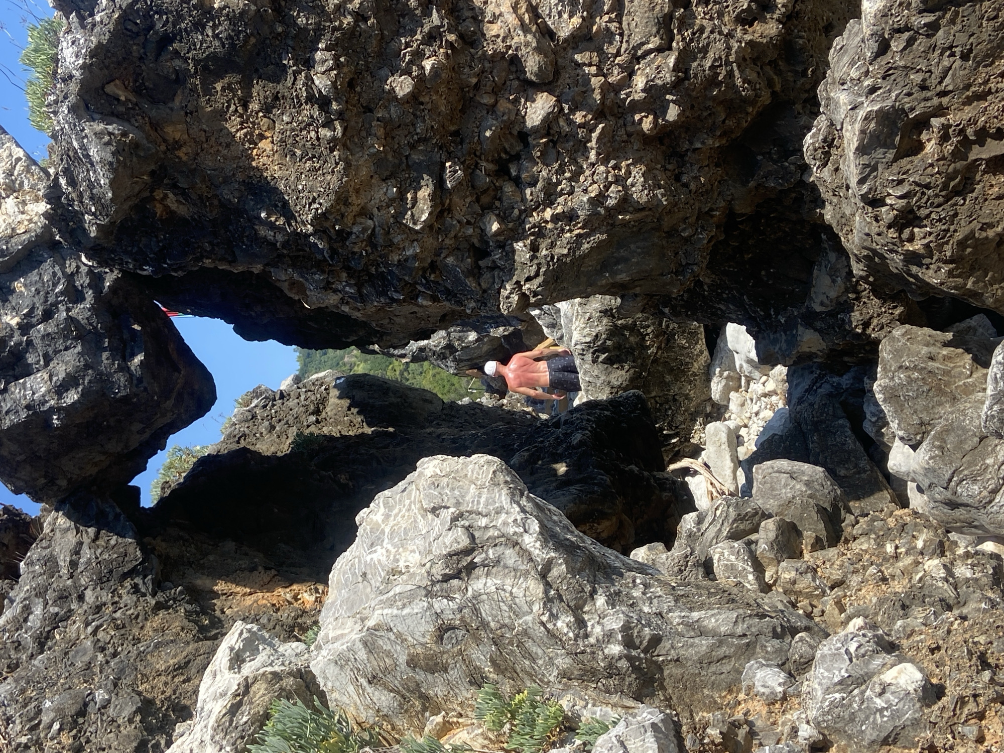 A person, with their back to the camera, stands partially obscured by a natural rock formation on a sunny day. The individual is wearing a white cap and dark-colored shorts, with their upper body bare. They are positioned within a rocky archway, with large, textured rocks dominating the foreground and framing the scene. Behind the person, lush green vegetation and a bright blue sky are visible, suggesting an outdoor, potentially coastal or mountainous, environment. The overall setting appears to be a rugged, natural landscape.