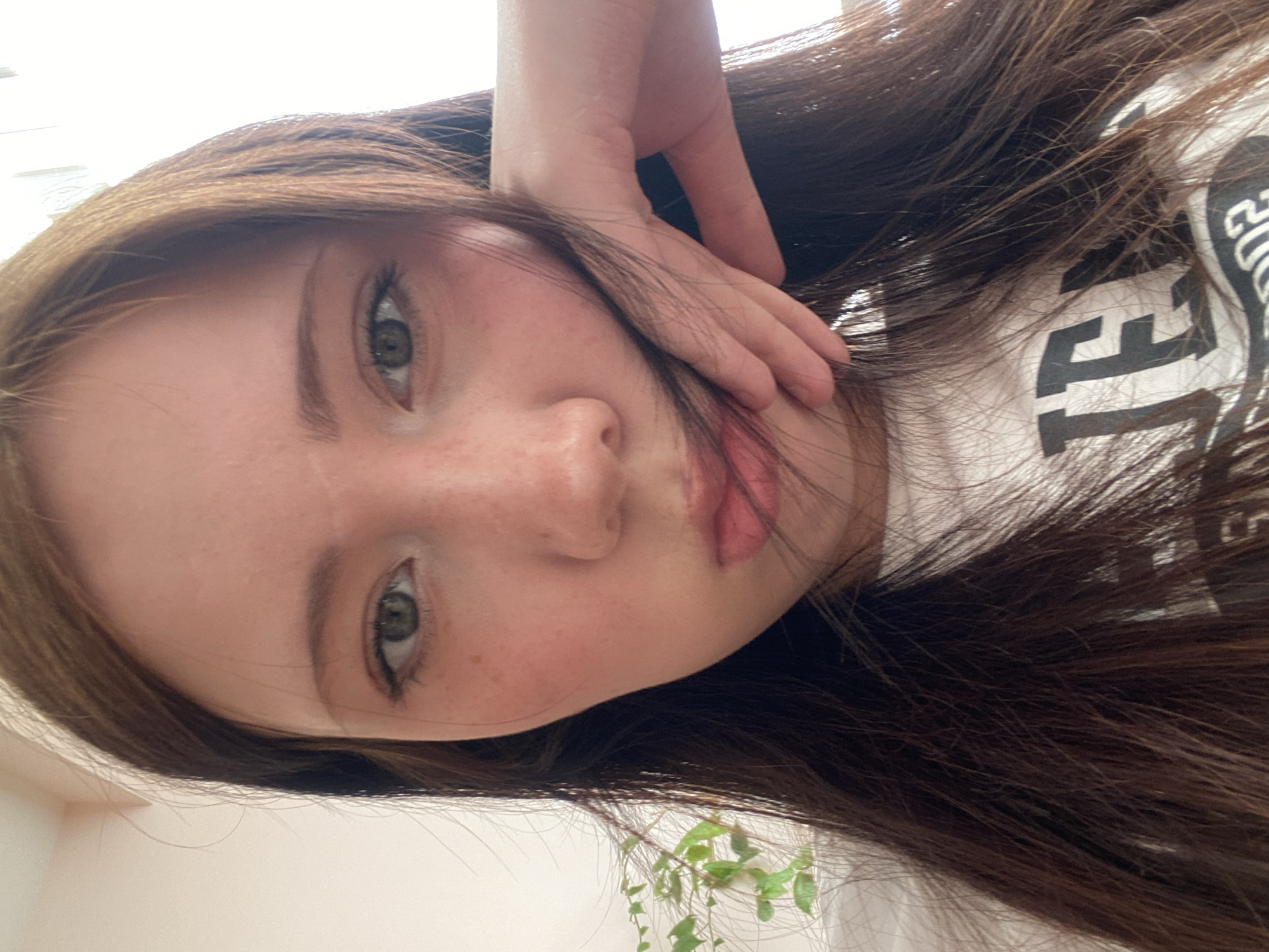 A close-up, angled selfie shows a young woman with light skin, freckles, and light brown eyes. Her dark brown hair is long and slightly disheveled, falling across her face and onto a white t-shirt with black lettering. She is resting her hand against her cheek. A small green plant is partially visible in the lower portion of the frame. The location is Unknown, Unknown.