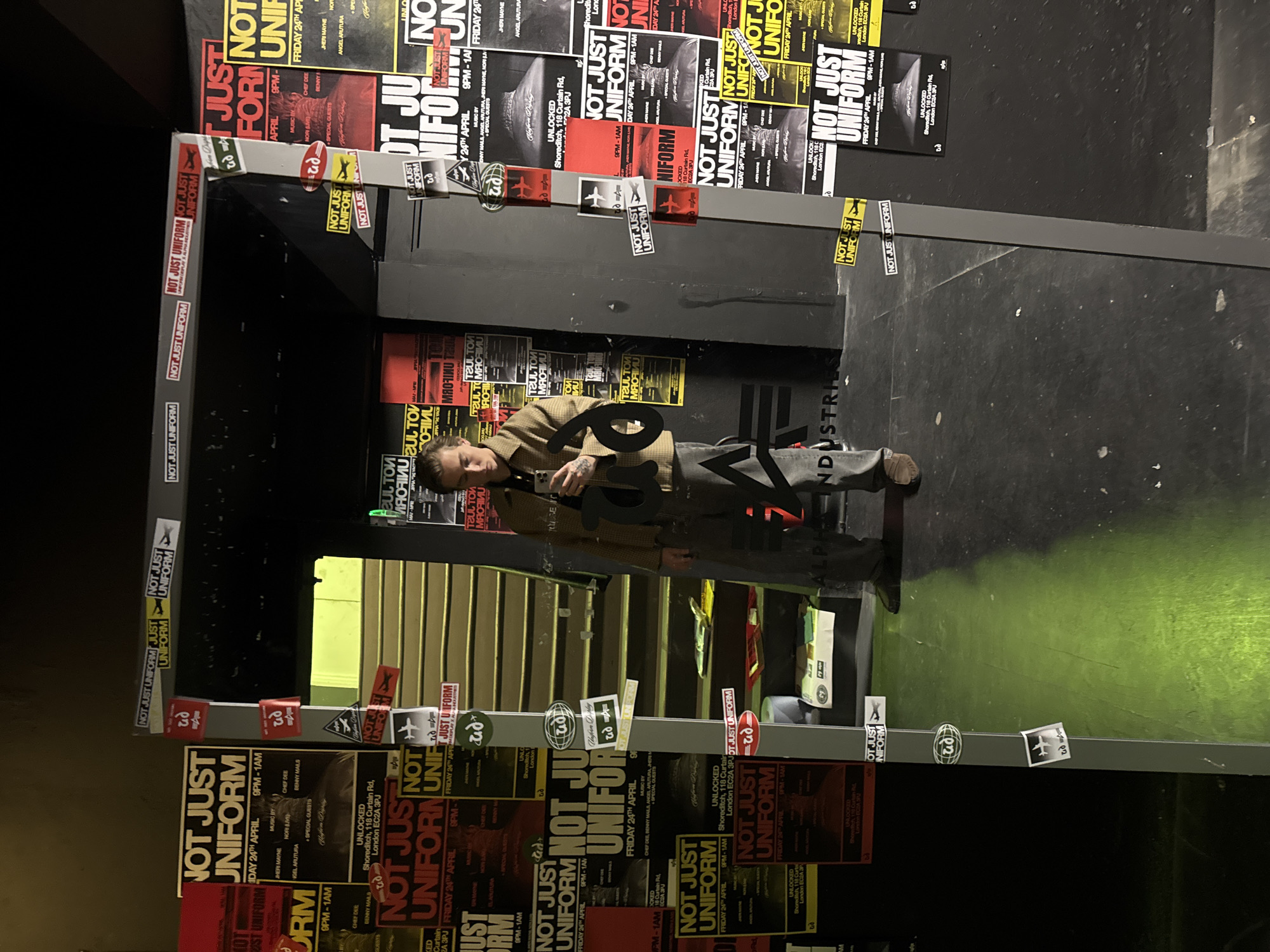 An indoor, vertically oriented shot captures a person standing within a doorway-like structure adorned with numerous event posters and stickers. The person, a male with short dark hair and visible arm tattoos, is wearing a plaid jacket and dark pants, holding two large, dark-colored cut-out numbers, "6" and "23." His expression is neutral.
The structure and surrounding walls are almost entirely covered in promotional material for an event titled "NOT JUST UNIFORM." These posters are predominantly in black, red, and yellow, announcing "FRIDAY 24TH APRIL" from "9PM - 1AM." The venue is specified as "UNLOCKED Shoreditch, 118 Curtain Rd, London EC2A 3PJ," confirming the London location context. Details about "MUSIC BY" various artists like JHERI MAYNE, NORI LIV, ANGEL ARUTURA, CHEF DEE, and DENNY MAILS are visible, along with mentions of "SPECIAL GUESTS."
Numerous stickers are affixed to the structure, including the "NOT JUST UNIFORM" logo, stylized "p2" (or "q2") emblems, an airplane icon, a globe icon, and the text "ad." Some partially visible or mirrored text on posters includes "TOUL TOM," "MRORINU," and "NDUSTRIE" (likely part of "INDUSTRY"). The background features dark walls and additional posters, suggesting a club or event space. The flooring is dark, with a greenish hue underfoot.