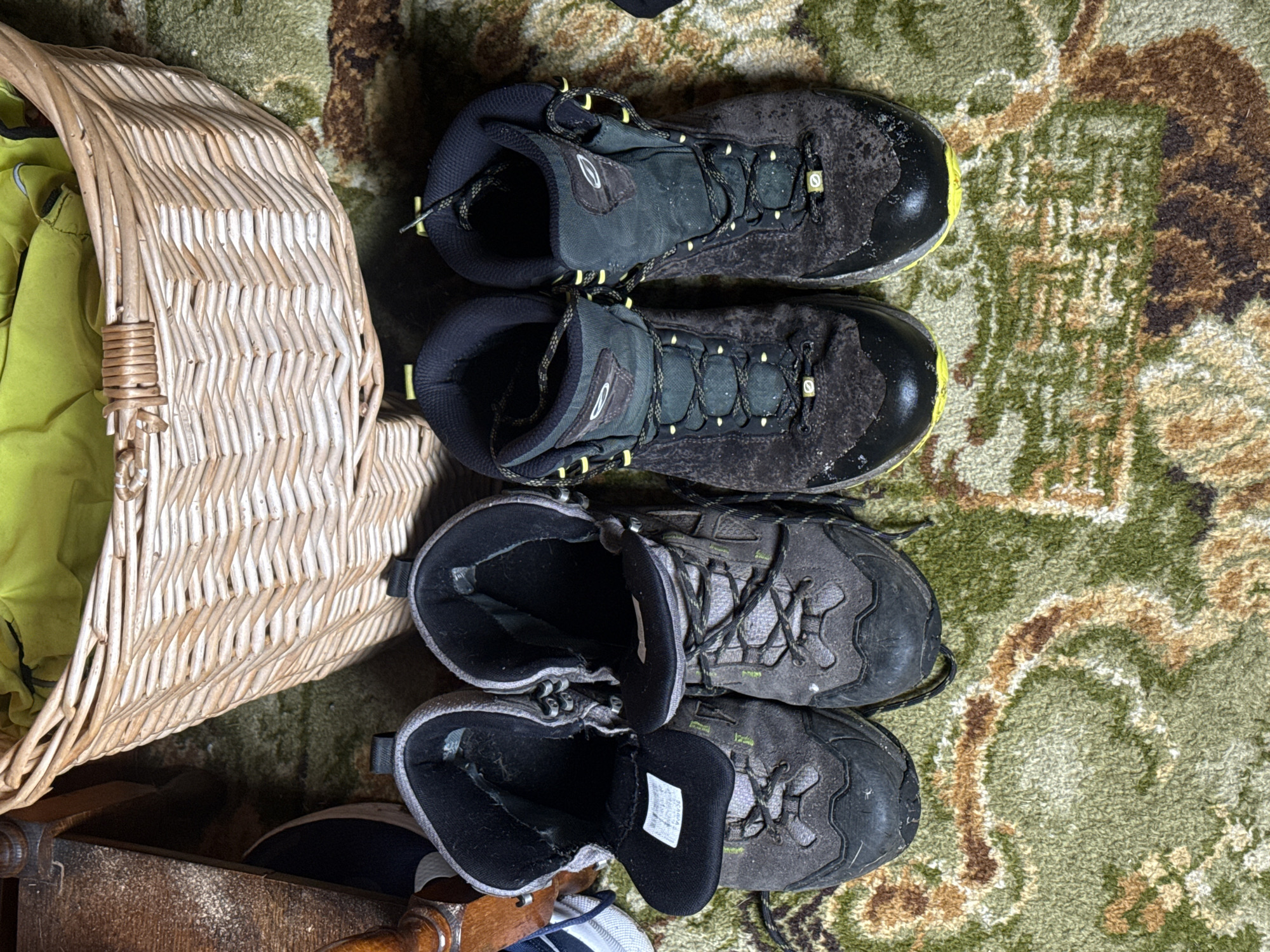 The scene depicts two pairs of hiking boots lying on a patterned rug. A wicker basket sits to the left, containing a bright green garment. The boots are worn and dusty, suggesting recent outdoor use. The rug features a mix of green and brown tones. The overall impression is one of equipment being stored or prepared for use in a cluttered domestic environment. No location or city can be identified. There are no people in the image.