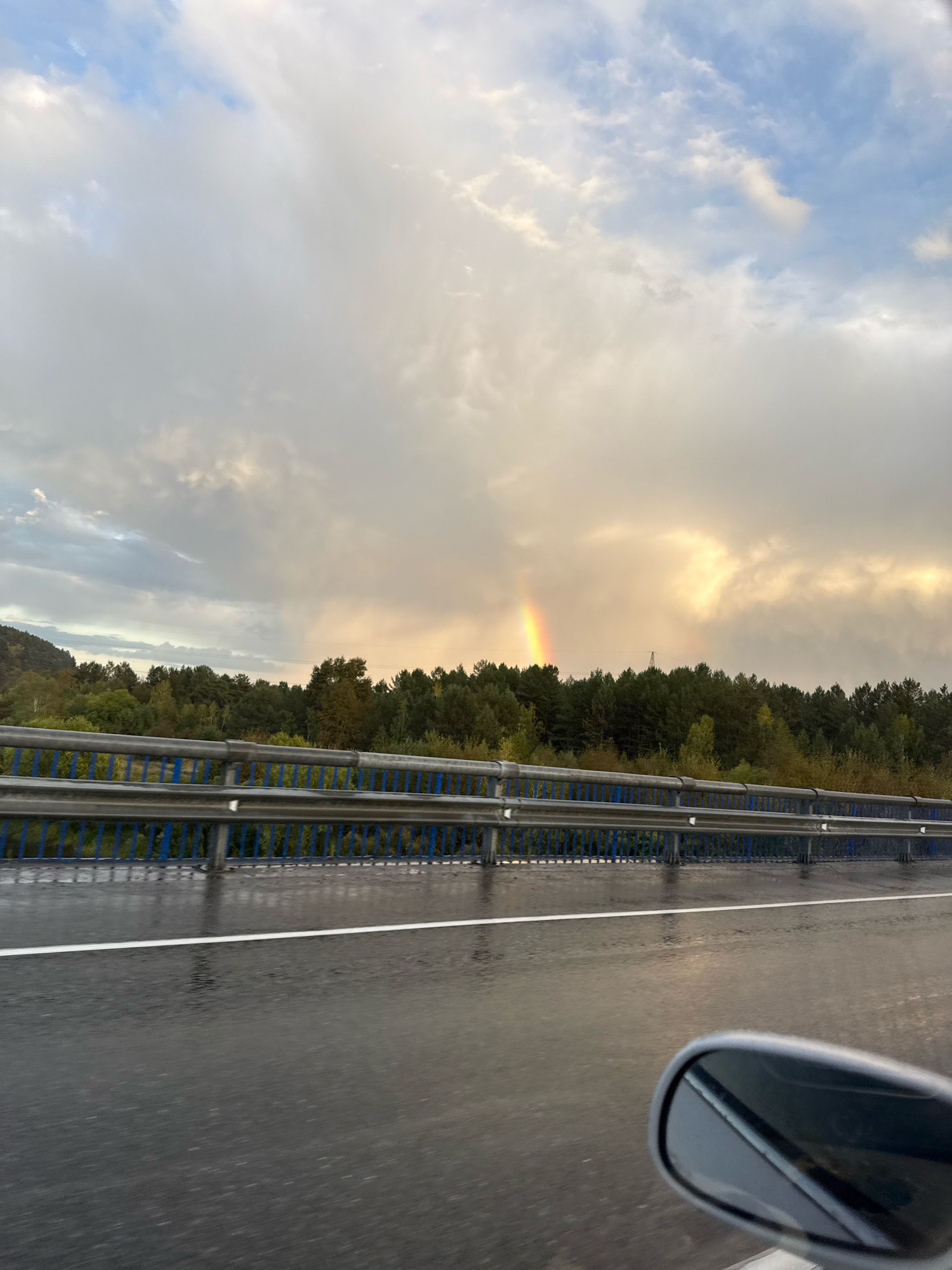 The image captures a scenic view from a vehicle traveling on a wet road, likely a highway, with a guardrail separating it from a treeline and a hilly landscape. The sky is dramatic, with a mix of dark, ominous clouds and patches of bright sunlight, suggesting recent or ongoing rain. A faint rainbow is visible in the sky above the trees. The road surface is reflective, indicating it is wet, and a white line marks the lane. A car's side mirror is visible in the lower right corner of the frame. The general scene depicts a natural landscape under a dynamic sky, possibly during a transition in weather. There are no people visible in the image, and the environment appears to be rural or on the outskirts of a settlement. The location of Pionersk, Russia is not evident from the image itself, as there are no landmarks or identifying features present.