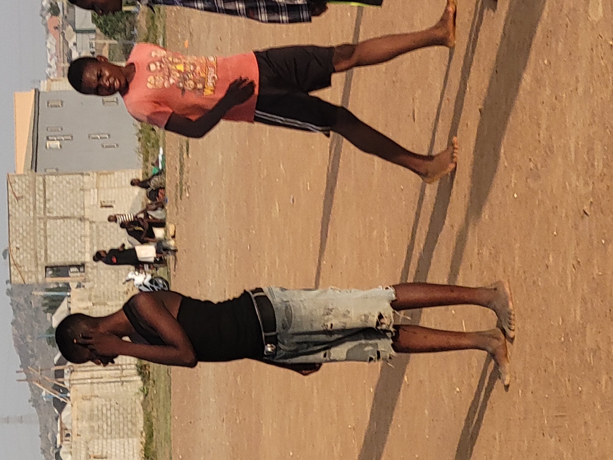 Two barefoot young males are observed on a dirt-covered ground surface. One male is wearing a dark-colored tank top, tattered jean shorts, and a belt, while the other male is wearing a light-colored t-shirt and dark-colored shorts. In the background, buildings and other people are present. This is the Ibo area of Nigeria.