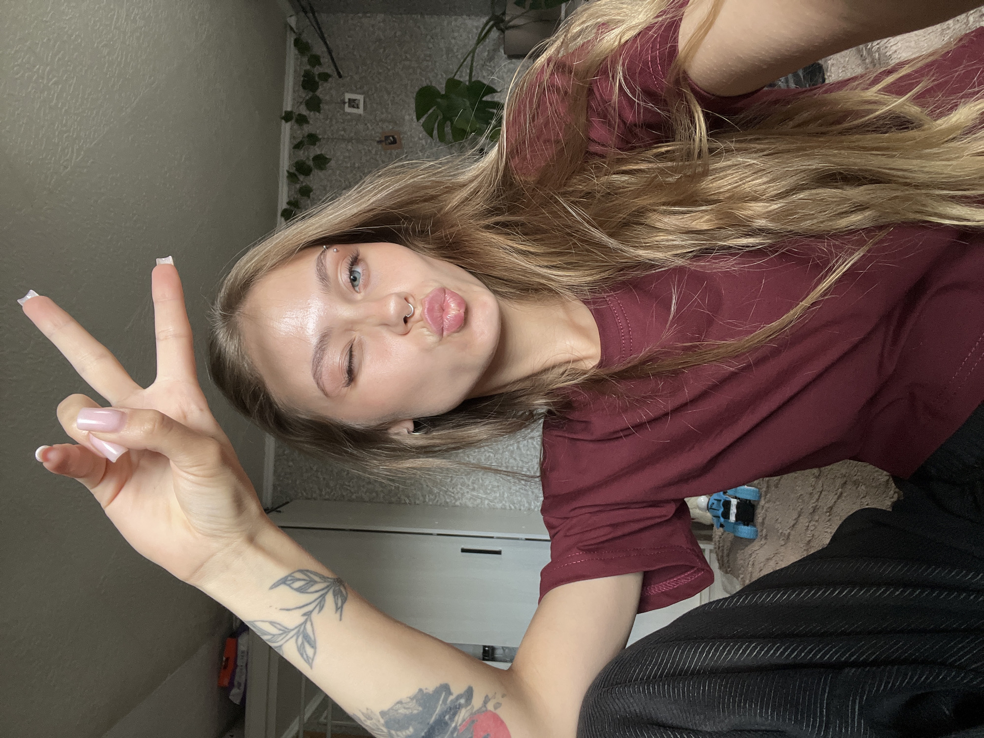 This rotated selfie captures a young woman posing playfully indoors. She has long, wavy blonde or light brown hair and is wearing a maroon t-shirt, with dark, subtly striped pants visible in the lower right. Her left hand is raised, forming a peace sign, revealing long fingernails with a light pink base and white tips.
Her face is angled towards the camera, with her right eye winking and her lips pursed in a "kiss" expression. She has a piercing with two studs on her right eyebrow and a ring piercing in her septum. Tattoos are visible on her right arm: a delicate leafy branch design on her inner wrist/forearm, and a larger, darker, abstract or possibly floral design on her upper arm.
The background shows a light-colored wall adorned with artificial ivy-like greenery. A large green leaf, likely from an indoor plant, is also visible. Part of a light-colored cabinet or furniture item is seen behind her left arm. The indoor setting appears to be a casual, personal space. The image conveys a cheerful and casual mood. No text is visible in the image.