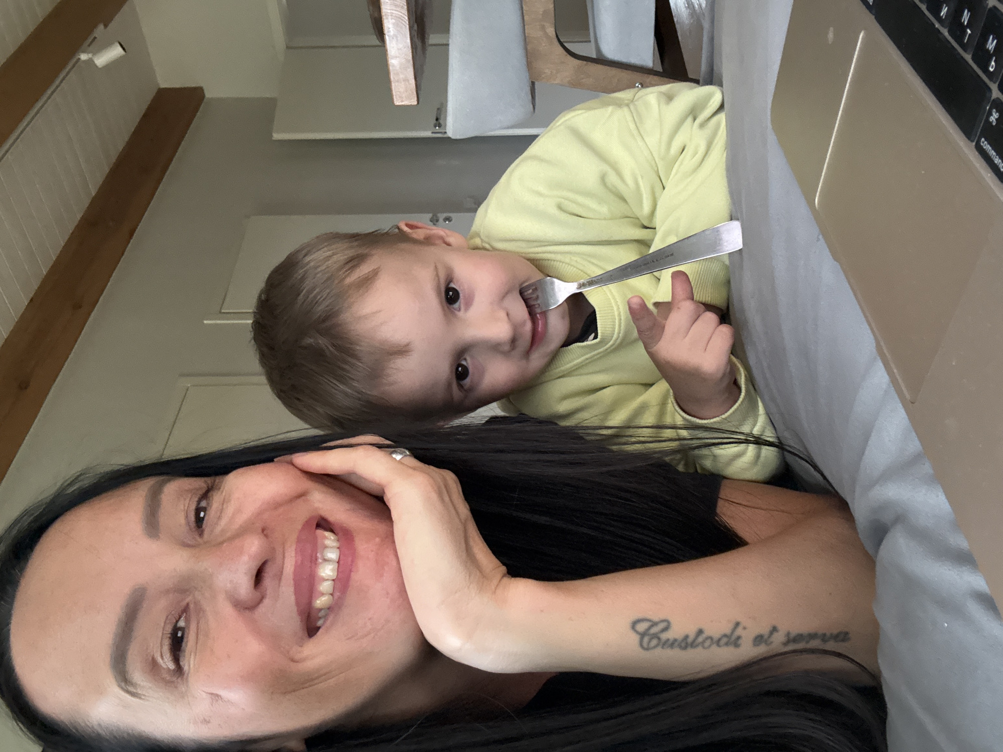 A woman with long dark hair and a tattoo on her arm is smiling at the camera, resting her chin on her hand. Beside her, a young boy with blond hair is looking at the camera and holding a fork to his mouth, pointing with his other hand. They are both on a gray surface, possibly a bed or couch. A laptop is visible in the foreground. The background appears to be an indoor setting with a light-colored wall and a wooden-beamed ceiling. The location is not specified to be in Chaikovsky, Russia.