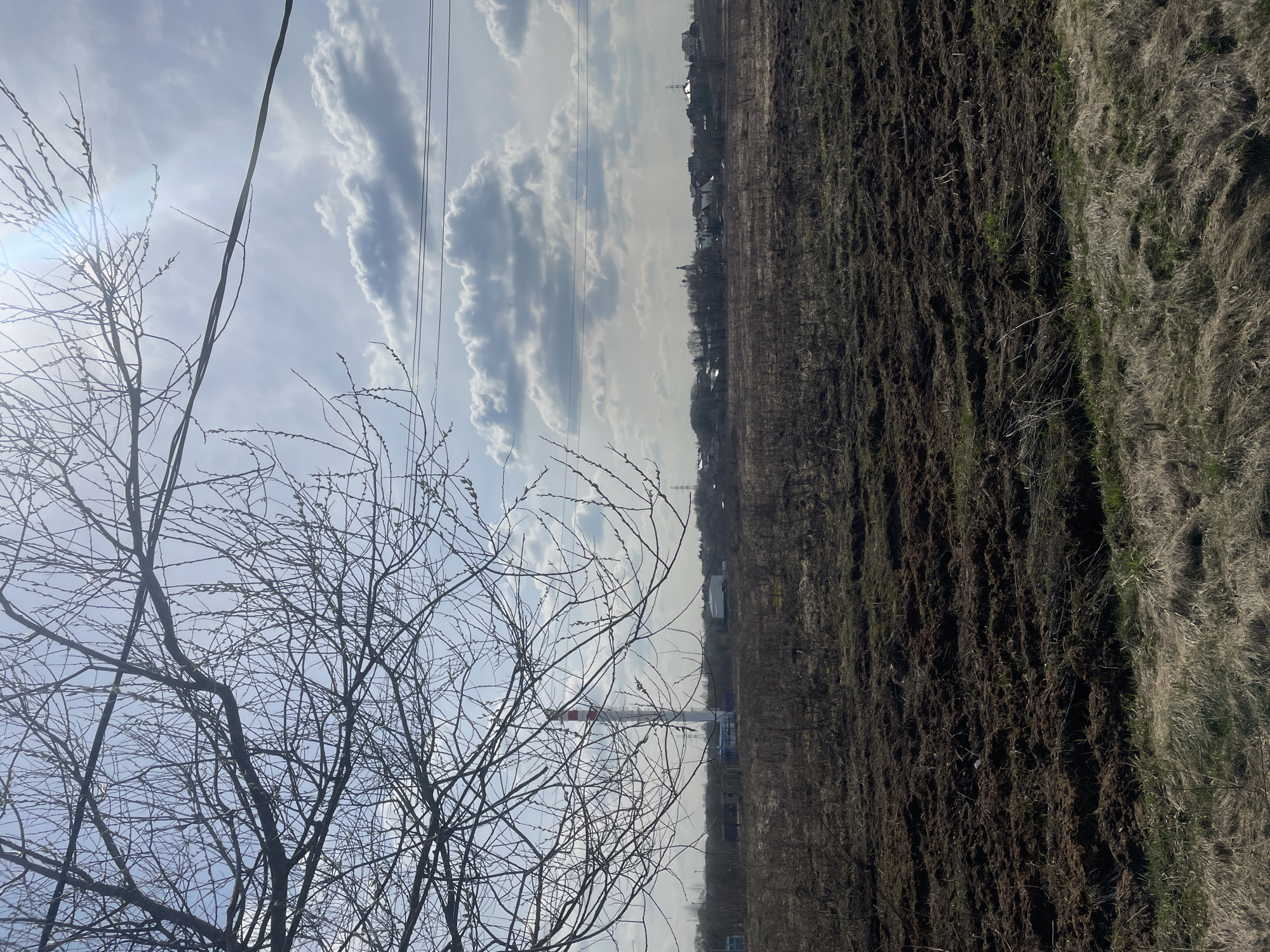 The image shows a field in Fabriki 1 maya, Russia, next to a bare tree in early spring. The sky is partly cloudy, with a mix of blue sky and white clouds. Power lines run across the sky. In the background, there are buildings, including a tall chimney with red and white stripes. The field is brown and appears to have been recently tilled. Patches of dead grass can be seen.