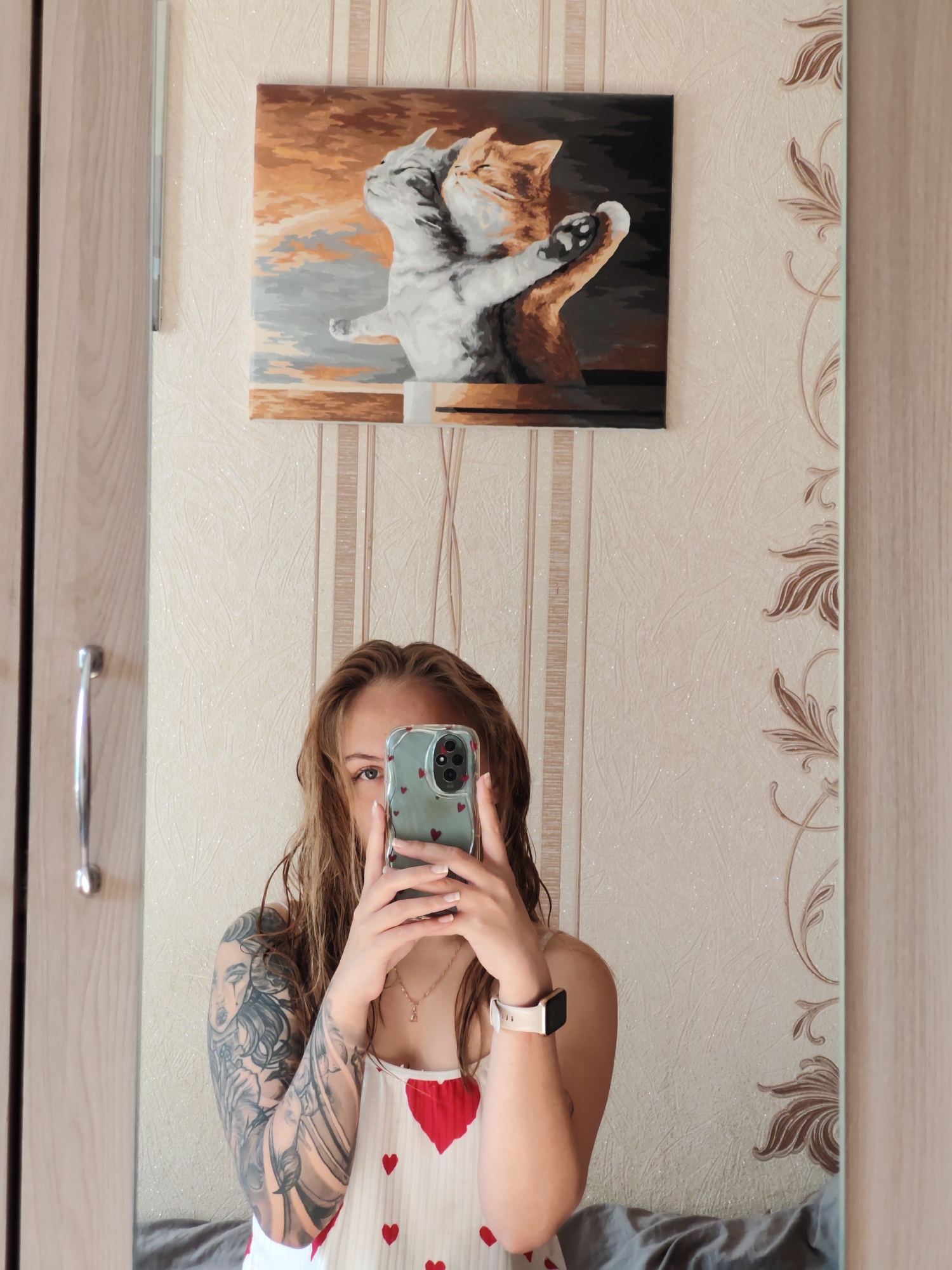 A person stands in front of a mirror in a room in Chaikovsky, Russia. The individual is holding a smartphone with a patterned case to take a photograph of themselves in the mirror. The person is wearing a white top with red heart patterns, a watch on their left wrist, and a necklace. The person has a tattoo covering their entire left arm. Behind the person and reflected in the mirror is a wall decorated with a textured, floral-patterned wallpaper. Above the individual is a framed painting depicting two cats in an embrace. To the left and right of the reflection are portions of a wooden wardrobe or closet. A silver handle is visible on the left. The bottom of the reflection shows a gray surface, possibly a bed.