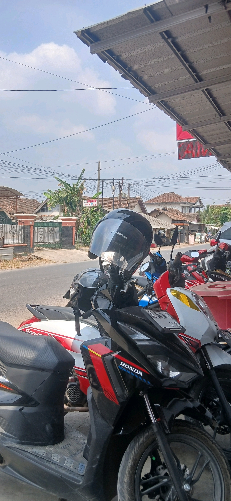 The image depicts an outdoor scene on a sunny day, likely in the city of Metro, Indonesia. In the foreground, several motorcycles are parked. The closest motorcycle is black with red and blue accents and the "HONDA" logo visible. A black helmet with a reflective visor is placed on its handlebars. To the left, another motorcycle is partially visible, featuring a white and red design with the word "Genio" on its side. Further back, more motorcycles, including a blue one, are parked.
The background shows a street with houses and buildings typical of a residential area. Power lines are visible crisscrossing the sky. A sign with text that appears to say "BARNIER" and "300 R" is mounted on a pole near a cluster of banana trees. The overall impression is a snapshot of everyday life in an Indonesian town.