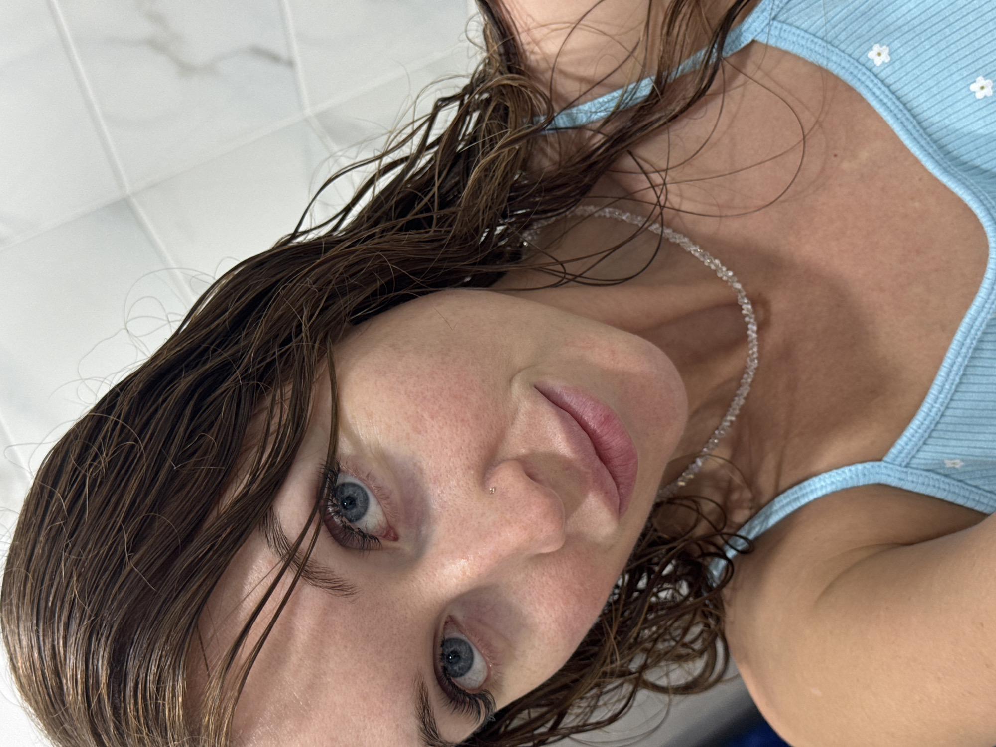 A close-up, low-angle view of a woman with wet brown hair and blue eyes, wearing a light blue top and a beaded necklace. A small stud is visible in her left nostril. The background appears to be a white tiled wall. The location is not discernible from the image.