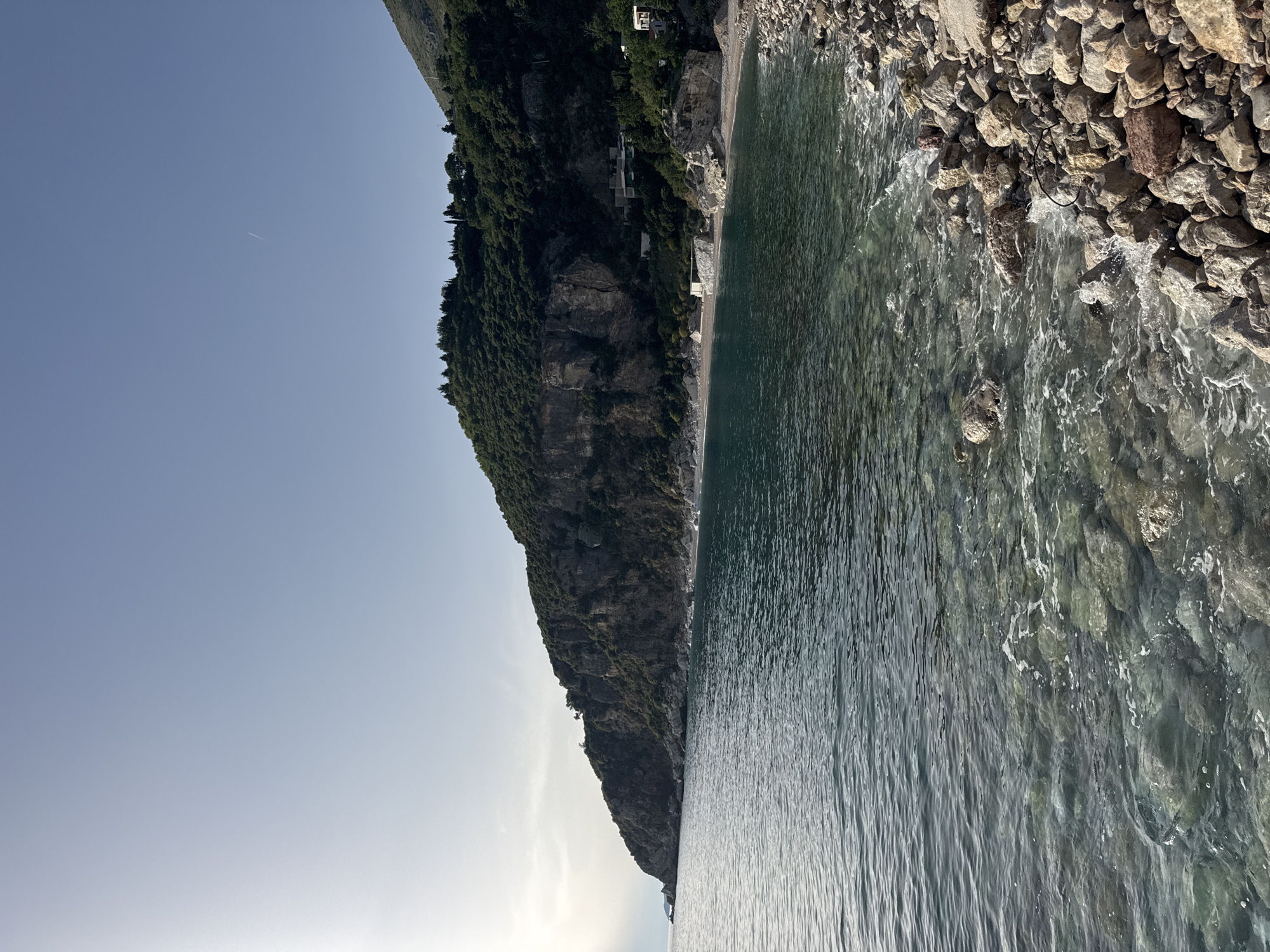 A picturesque view in Perazića Do, Montenegro features a steep, verdant mountain rising from the shoreline. The clear turquoise waters lap against a rocky beach, with the horizon meeting a pale blue sky. The sunlight illuminates the scene, creating a serene and tranquil atmosphere. Buildings are nestled into the mountainside.