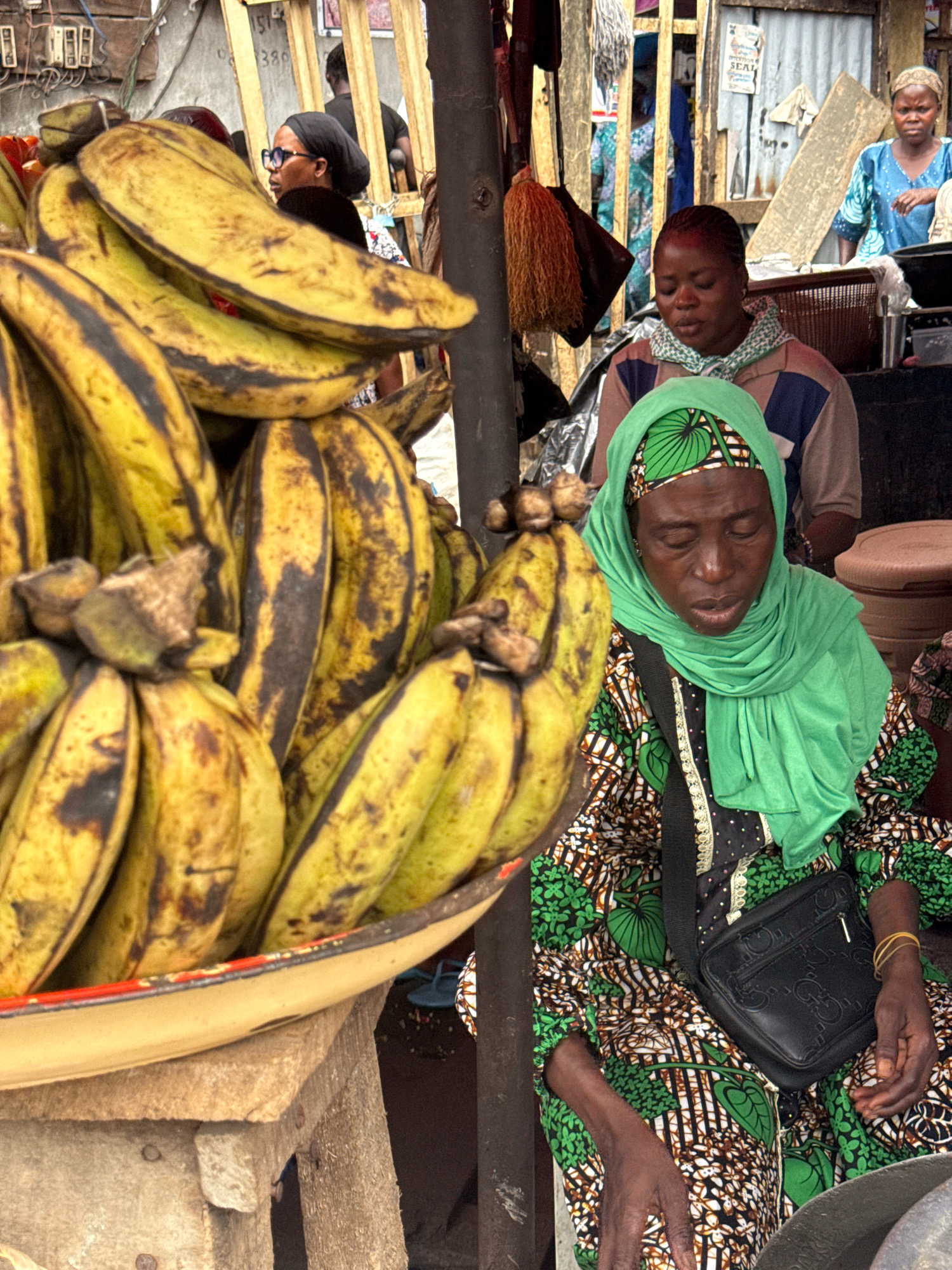 In a street market in Lekki, Nigeria, a woman wearing a green headscarf and a patterned dress sits with her eyes closed, her head bowed. A black purse with a gold zipper is slung across her chest. To her left, a large pile of ripe plantains sits in a yellow bowl. In the background, other market stalls and people are visible, including a woman in a blue dress to the right and another person partially obscured by the plantains to the left. The market appears to be bustling, with various structures and wares present.