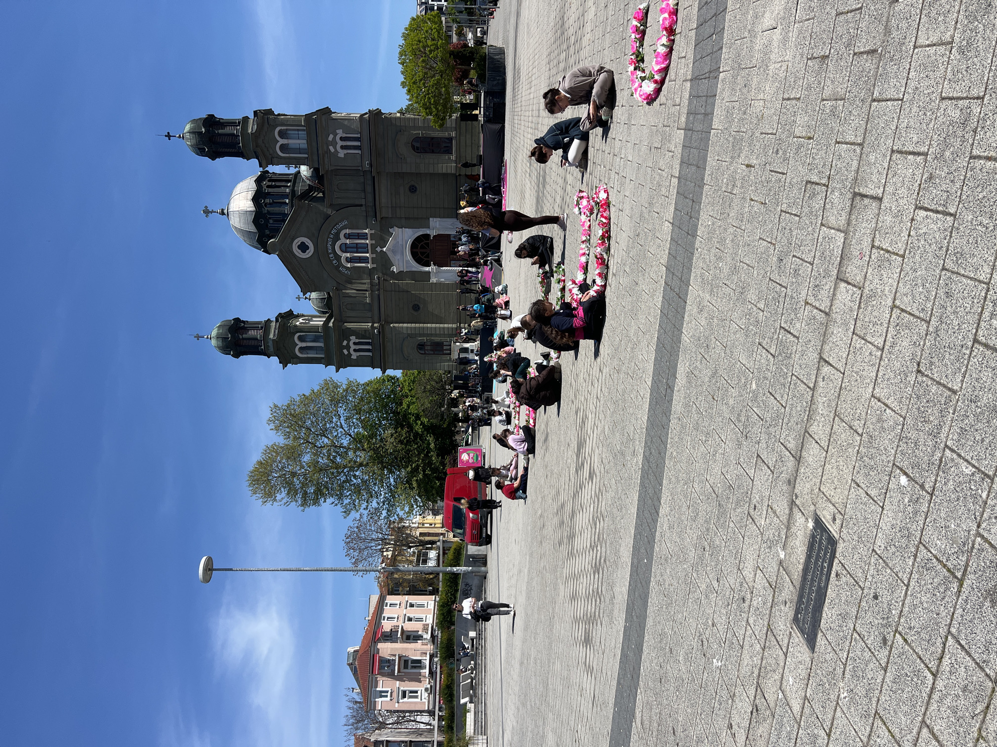 The image shows the Cathedral of Saint Cyril and Methodius in Burgas, Bulgaria, from an outdoor perspective. A group of people are near a flower arrangement on a paved area. The weather appears clear. A streetlight and buildings can be seen in the background.