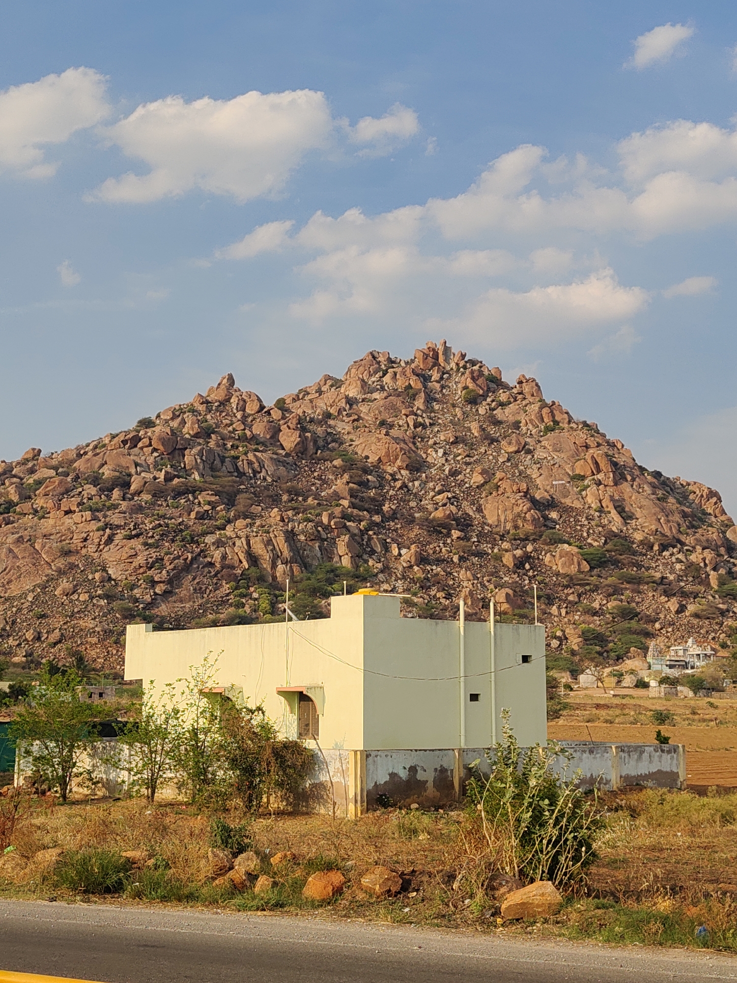 The image shows a daytime view in Sanjivapuram, India, featuring a beige building in the foreground, with a rocky hill in the background. The building is a single story with a flat roof and minimal architectural details. Vegetation surrounds the base of the building. A low, weathered concrete wall extends around a portion of the building's perimeter.
The hill behind the building is covered with large, reddish-brown rocks and sparse vegetation. In the distance, another structure resembling a building is visible. The sky is blue with scattered white clouds. A road runs along the bottom of the frame, and the ground in the foreground is dry and brown.
