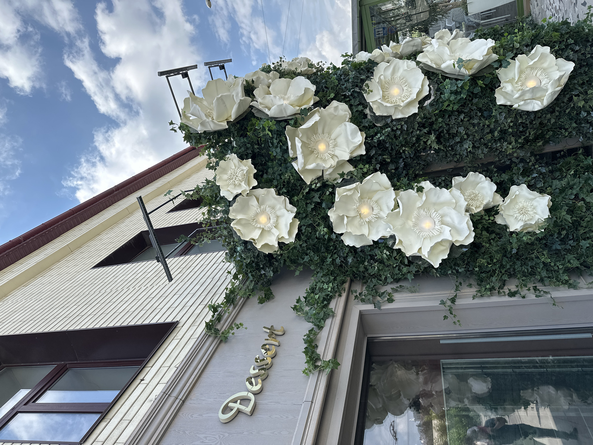 The image shows a facade of a building with a sign that reads "Dessert". The facade is decorated with large, white paper flowers and ivy. The building appears to be located in a city, with a blue sky and clouds visible in the background. A reflection in the window shows a person holding a phone. Based on the context of previous images, this location is in Yekaterinburg, Russia.
