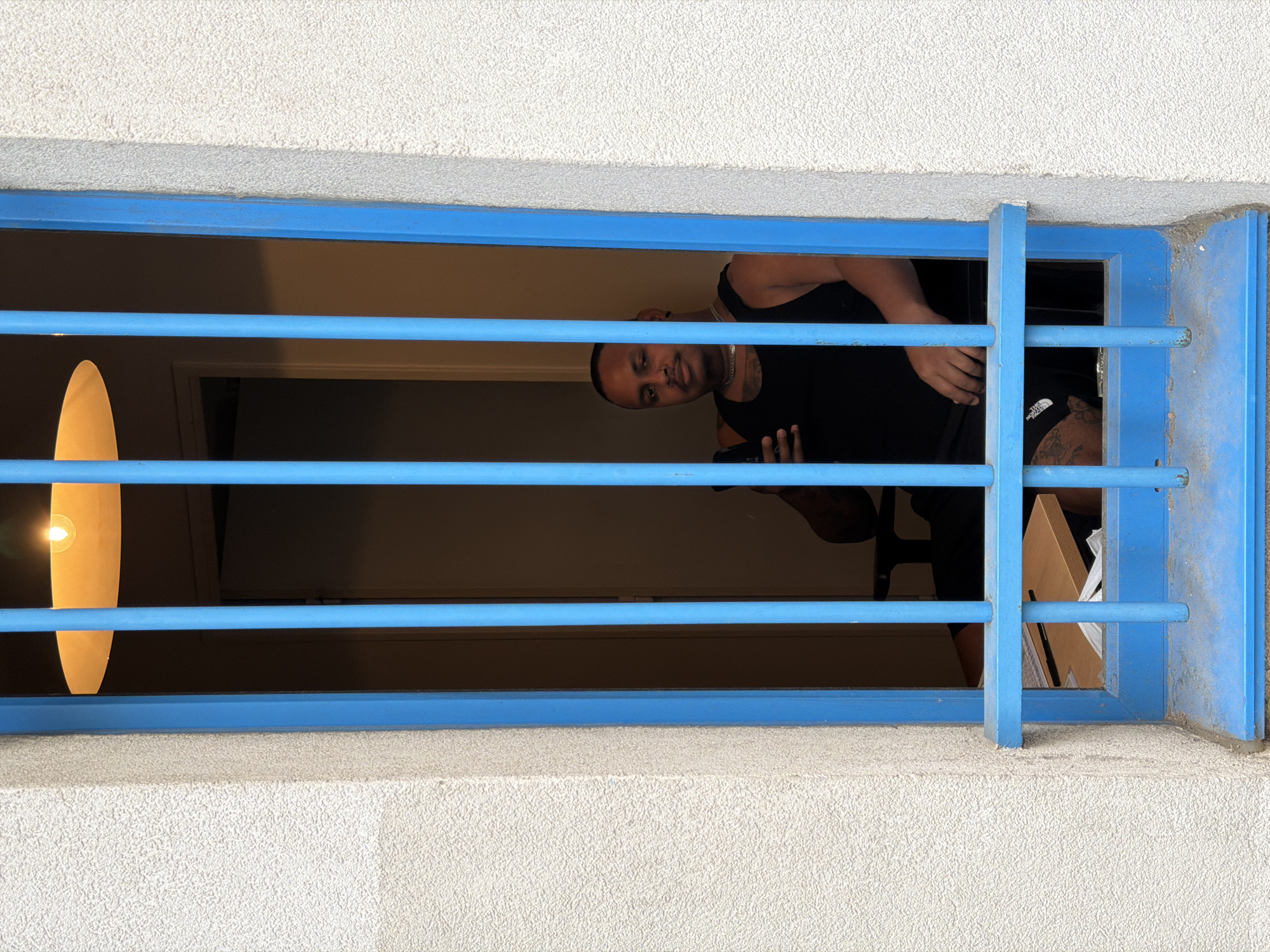 An adult male wearing a black tank top and jewelry is seen looking towards the camera through a blue barred window. He is seated on a chair, holding a mobile phone in his right hand. The background is dimly lit, with a desk visible to the left and a light fixture overhead. The exterior of the building features a textured, light-colored wall surrounding the window. The overall scene suggests an interior space viewed from the outside, with the blue bars serving as a visual obstruction. There is no specific indication of the city of Paris, France.