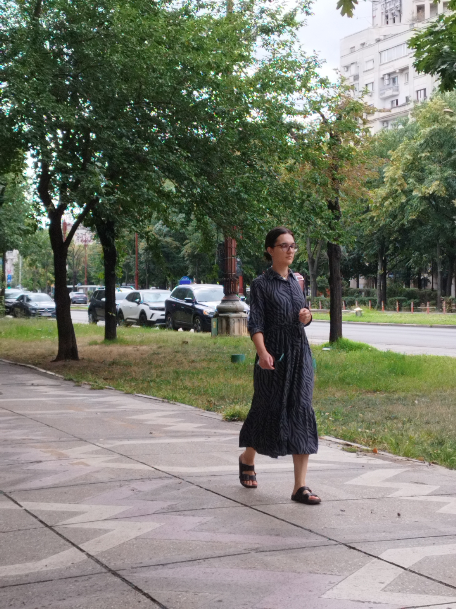 A woman with dark hair and glasses, wearing a dark, patterned, ankle-length dress and black sandals, walks along a concrete sidewalk next to a street in Bucharest, Romania. The sidewalk has a geometric pattern. To the right of the sidewalk is a grassy area and then a street with several cars parked and driving. Behind the street are trees and tall buildings, some of which appear to be apartment buildings. The trees are green with some autumnal colors visible. The sky is overcast. The woman is looking forward and to her left, holding a dark object in her right hand. She is carrying a brown bag over her right shoulder. The scene appears to be an ordinary day on a city street.