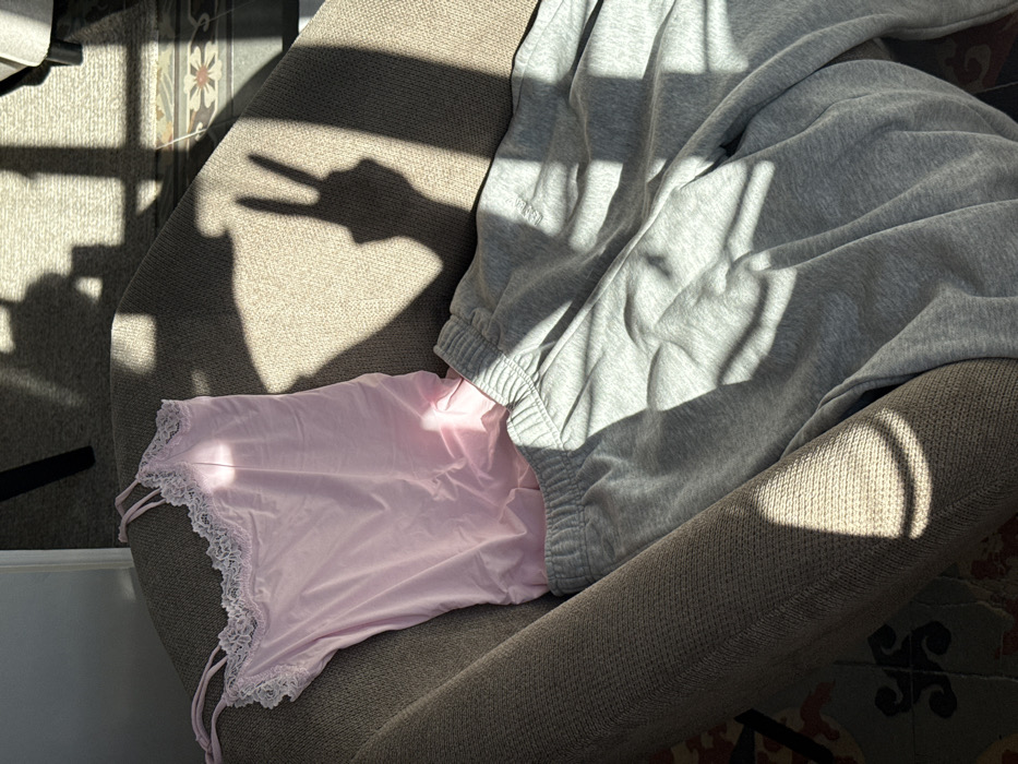 The image depicts an indoor scene within a room in Barcelona, Spain. A light brown, textured upholstered armchair is positioned centrally. Draped over the armchair are a light pink camisole with white lace trim along the neckline and spaghetti straps, and a pair of grey elastic-waisted sweatpants. Strong sunlight from an external source, likely a window, illuminates the scene from the right, casting prominent shadows. A distinct shadow of a human hand forming a peace sign gesture is visible on the chair's upholstery, to the right of the clothing. Linear shadows from a window frame or blinds also traverse the chair and the surrounding area. A portion of a tiled floor with a decorative, possibly floral or geometric, pattern characteristic of Catalan architecture is visible beneath and to the left of the chair. Indistinct furniture legs and further shadows are visible in the background.