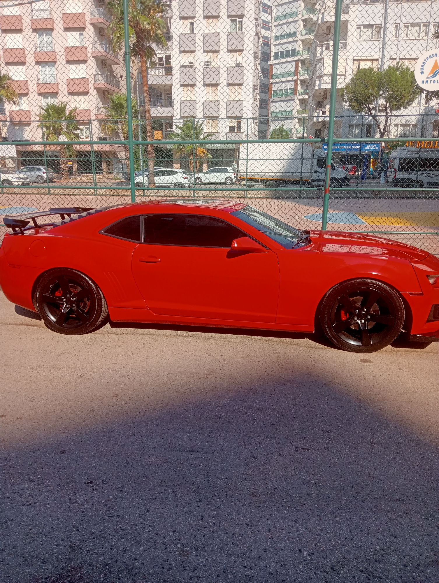 A bright red Chevrolet Camaro coupe is parked on an asphalt road in front of a chain-link fence. The car has black rims, a black rear spoiler, and tinted windows. Behind the fence, there is a multi-story building with a facade of beige and light grey squares, and balconies. Palm trees and other greenery are visible between the fence and the building. A white truck with "JET MELLE" written on its side is parked behind the fence, and a white van is parked next to it. The ground is a mix of asphalt and a lighter colored surface with yellow and blue markings, possibly a sports court. The scene appears to be in Antalya, Turkey, indicated by the "MURATPASA ANTALYA" text on a circular sign visible in the upper right portion of the image. Sunlight casts a shadow of the fence onto the road and car.