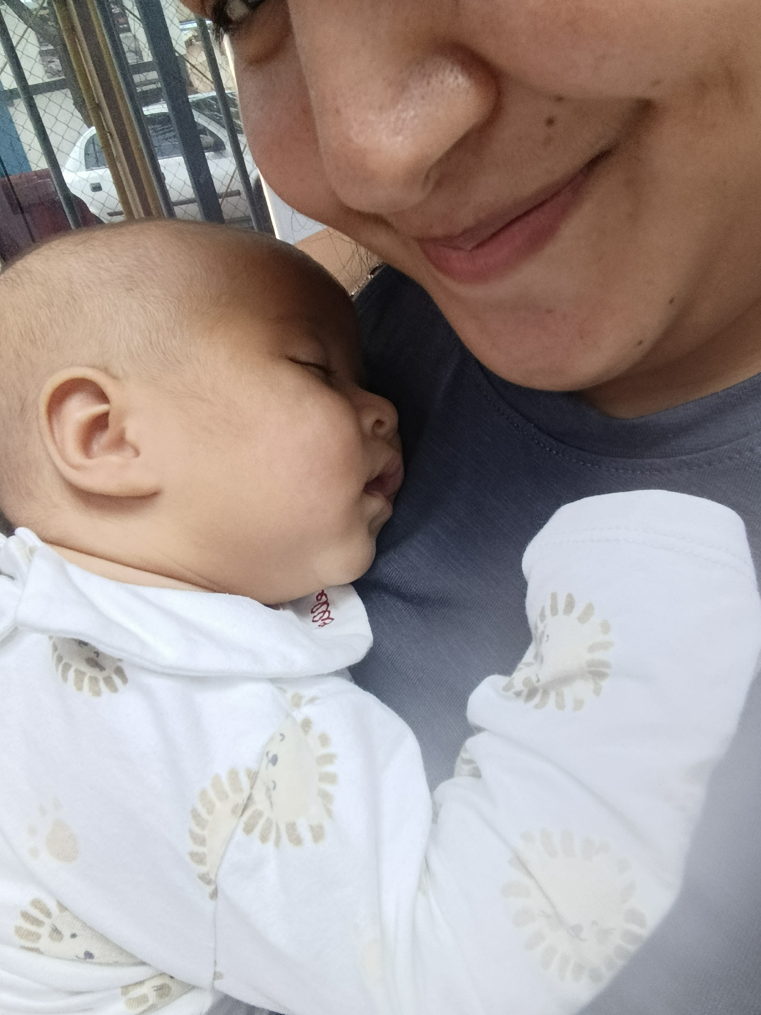 This close-up, self-taken photograph shows a person holding a sleeping baby. The baby is nestled against the person's chest, with its head turned to the side and eyes closed. The baby is wearing a white onesie with a pattern of stylized lion faces and paw prints. The person's face is partially visible, with their chin and mouth in focus, smiling gently. The background shows a blurred view of a white car and fencing, suggesting an outdoor setting, possibly a residential area. The lighting indicates it is daytime, with soft natural light. The overall mood is tender and affectionate, capturing a moment of quiet intimacy between the person and the baby. No text is visible in the image.
