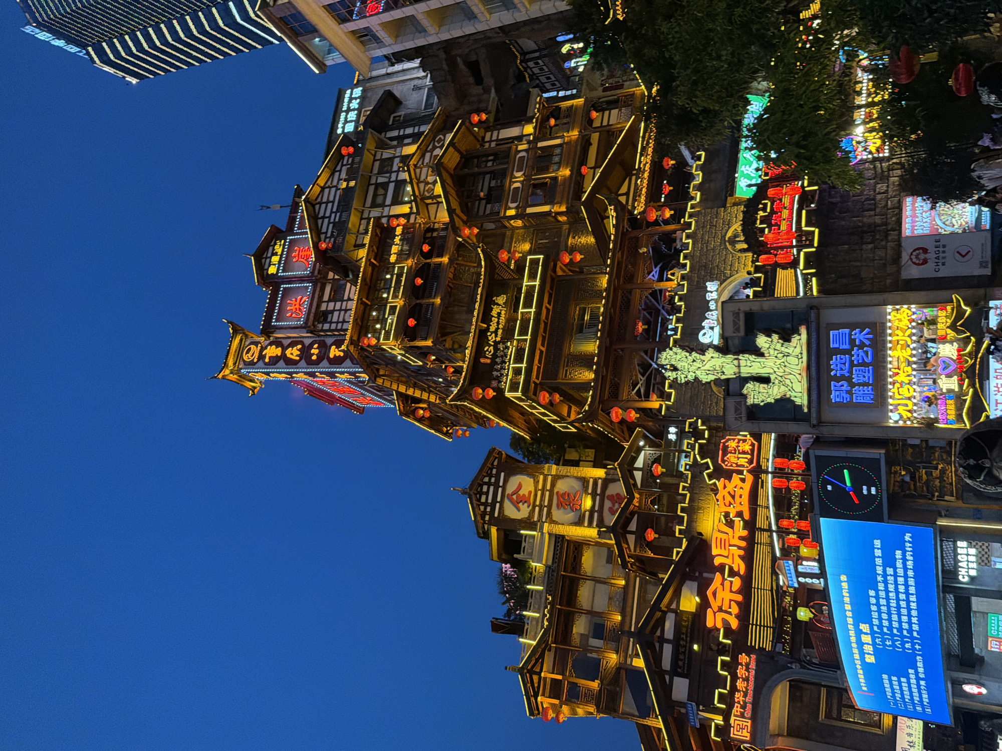 In Yuzhong District, China, at night, a multi-story building with traditional Chinese architecture is illuminated by numerous yellow lights and red lanterns. The building features tiered roofs and intricate wooden detailing. A modern skyscraper with a striped facade rises in the background. Several signs with Chinese characters are visible, some lit with neon lights. A digital screen displays text and a clock face. In the foreground, the tops of people's heads and shoulders are visible, suggesting a street-level view. The sky is a deep blue, indicating dusk or night.