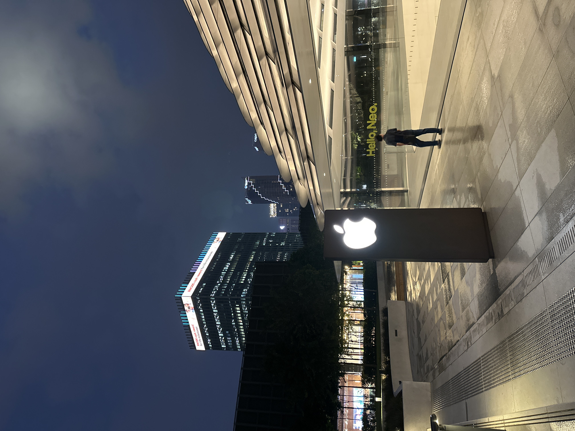 The image captures a modern urban scene at night, likely in Kuala Lumpur, Malaysia, given the context. The sky is dark blue, indicating late evening or nighttime. The ground, a mix of light and dark grey paving stones, appears wet, suggesting recent rain or standing water. Drainage grates and tactile paving lines are visible on the ground.
In the foreground, a brightly illuminated white Apple logo is displayed on a dark rectangular sign, standing vertically. To its right, a person is seen from behind, walking away from the camera on the wet pavement. They wear a dark top and blue jeans, casting a subtle reflection on the damp surface.
A large, contemporary building with a distinctive wavy, ribbed facade extends from the left side of the frame. Adjacent to it, and reflecting the night lights, is a glass-fronted building. On this glass facade, the text "Hello, Neo." is clearly visible, illuminated in yellow.
In the background, several tall, illuminated skyscrapers punctuate the skyline. One prominent building displays a large, brightly lit sign with the repeated phrase "RM500 cash reward for life milestones" on its upper levels. Another distant building shows the word "capri". The overall setting evokes a vibrant commercial district with sleek, modern architecture.
