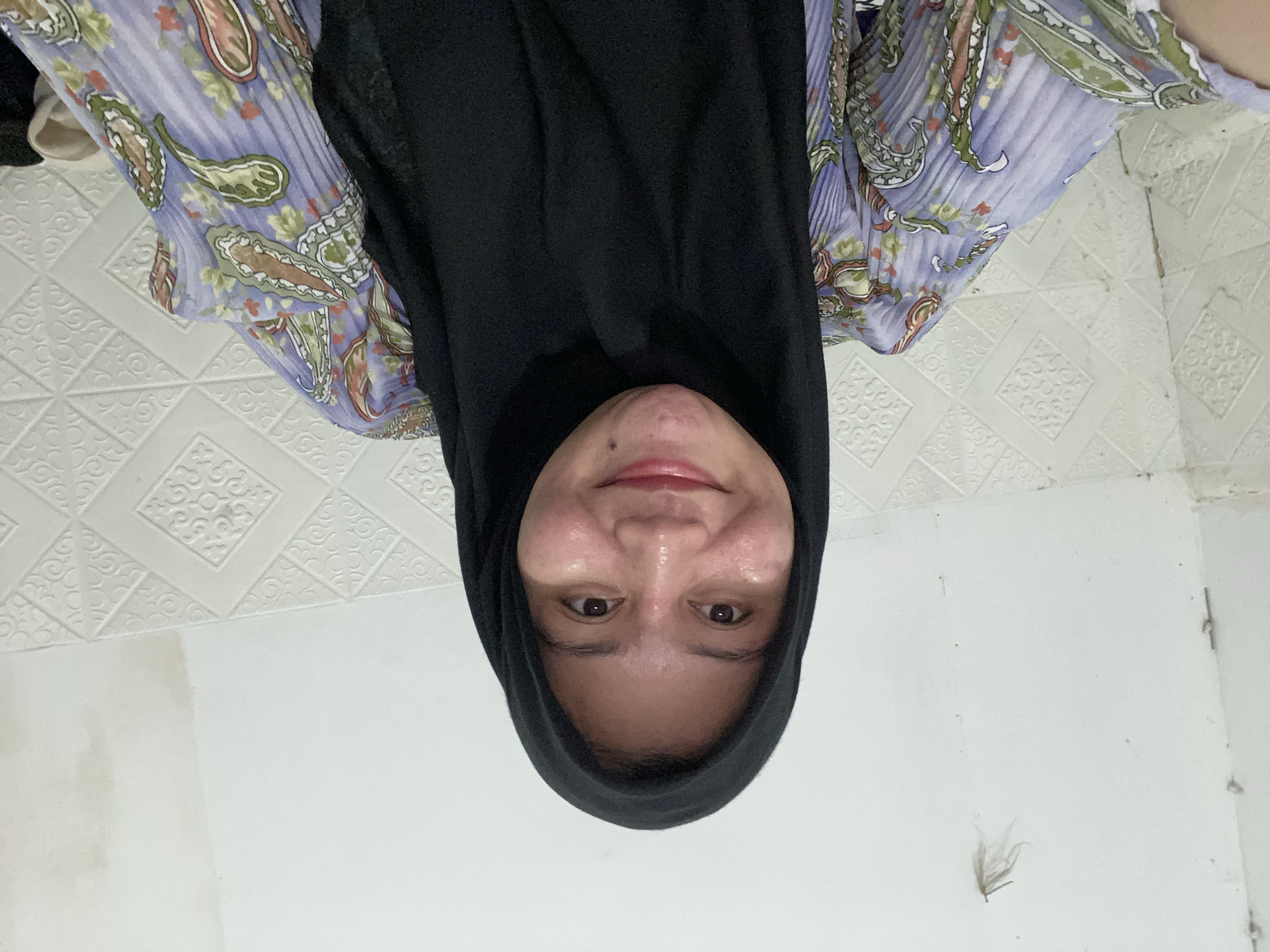 A selfie taken by a young woman wearing a black hijab and a light purple patterned blouse. She is smiling and looking directly at the camera. The background appears to be an interior wall with a textured pattern. There is a faint, small, dried leaf-like object on the white wall to the left of the woman's head. The scene suggests a casual indoor setting, possibly a home in Banjarbaru, Indonesia, based on the provided context. The lighting is soft, and the image quality is average, consistent with a selfie taken with a mobile device.