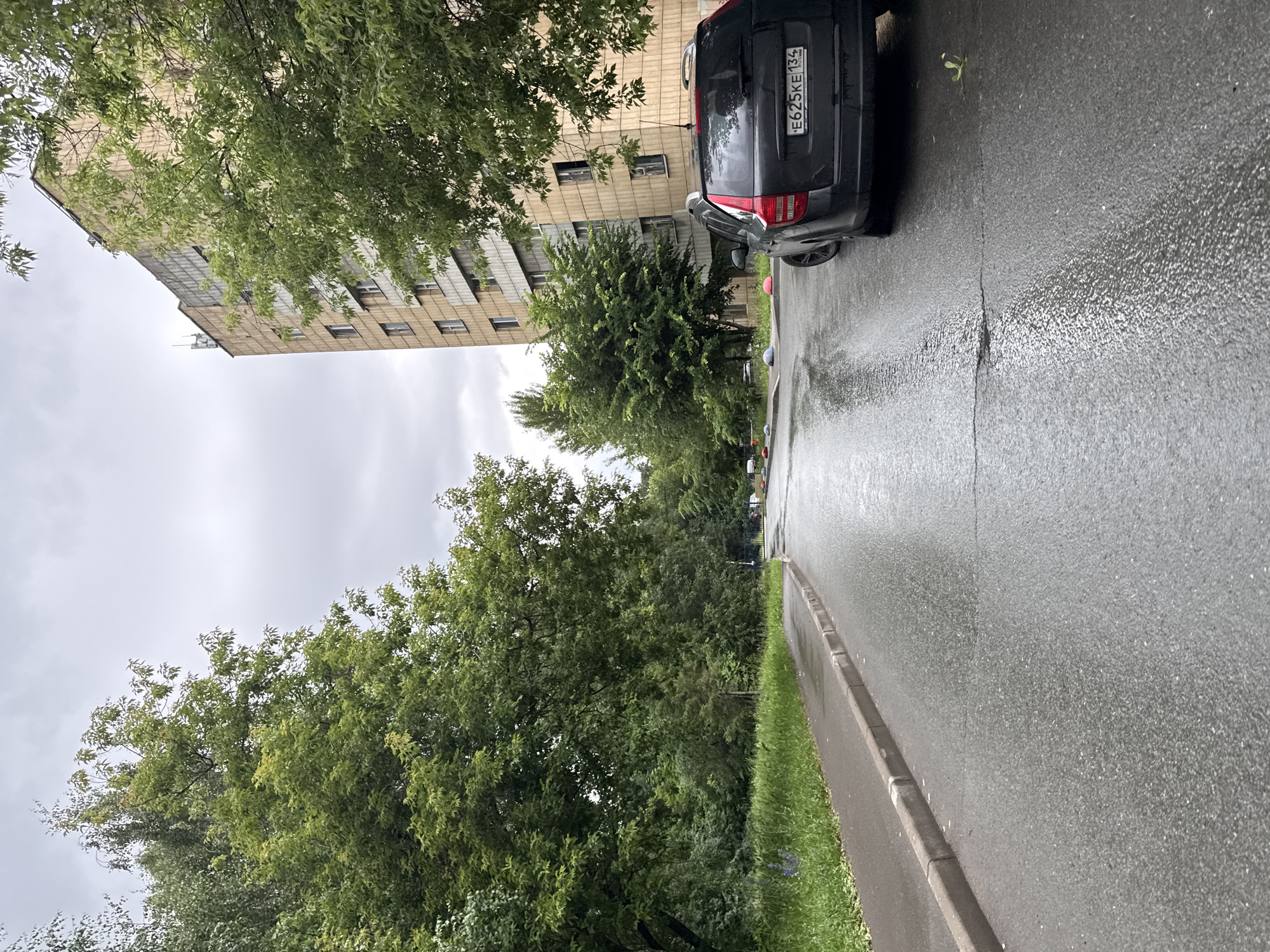 The image depicts a wet asphalt road in what appears to be a residential area in Kazan, Russia. The road is flanked by a sidewalk on the left and a line of trees on the left and right. In the background, a multi-story apartment building with a light-colored facade is visible, partially obscured by foliage. A dark-colored car, possibly a station wagon, is parked on the right side of the road, facing away from the viewer. The car's license plate is visible and reads "E625KE 134". The sky is overcast, suggesting recent or ongoing rain. The overall scene is one of a typical urban environment after a rainfall, with a focus on the road, the parked vehicle, the apartment building, and the surrounding greenery.