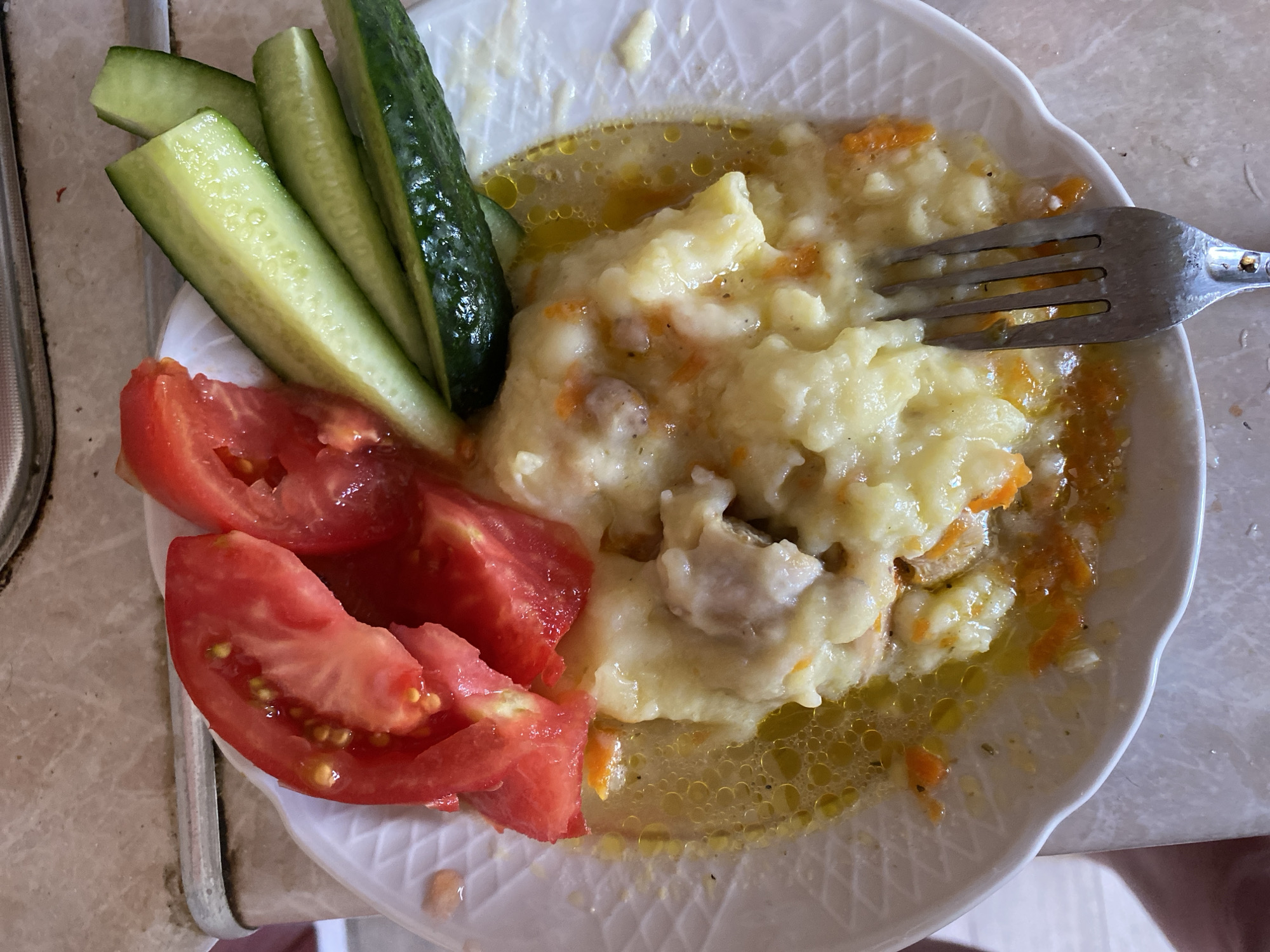 A close-up, overhead shot shows a plate of food with a fork on the right side. The plate is white with a scalloped edge and contains a portion of mashed potatoes with visible chunks of meat and carrots in a broth. Slices of tomato and cucumber are arranged on the upper left of the plate. The food appears to be on a table with a speckled beige surface. The image is set in the city of Molodizhne, Ukraine.