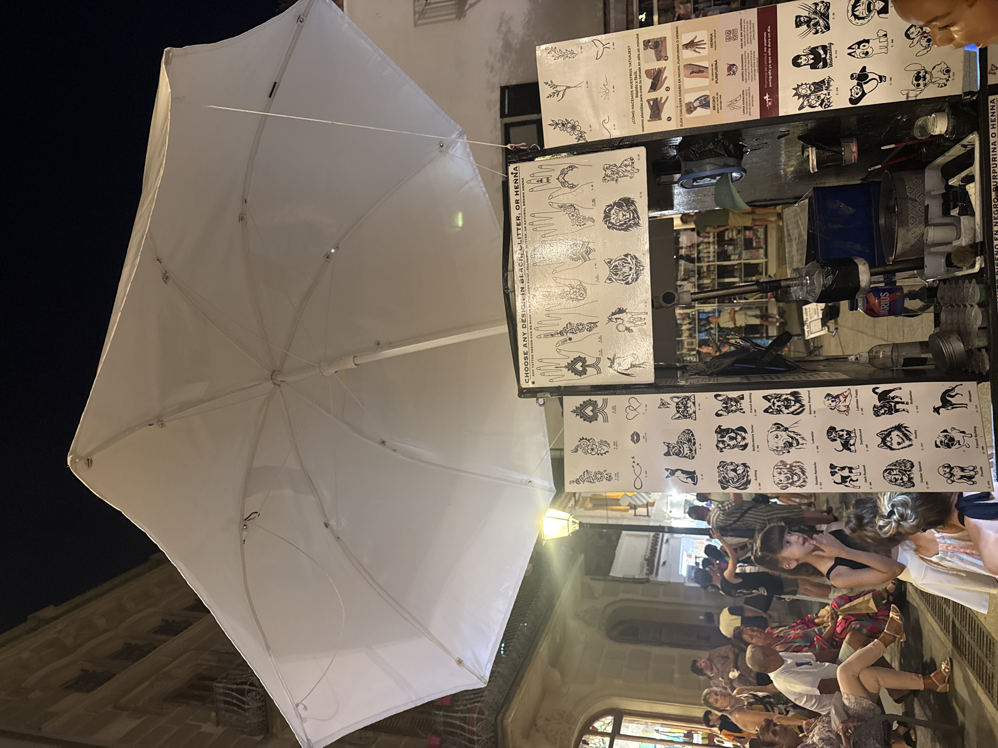 This image captures a lively evening scene in Tossa de Mar, Spain, likely a popular tourist area. The foreground is dominated by a large, off-white umbrella, casting a soft glow from a nearby streetlamp onto the street below. The umbrella is angled, revealing a glimpse of its structure.
Behind the umbrella, a stall is set up, displaying various tattoo designs. Several sheets of temporary tattoos are visible, featuring intricate patterns, popular cartoon characters like Wednesday Addams, and a variety of dog breeds, including a Cocker Spaniel, Poodle, and Whippet. Text in Spanish and English advertises the services: "CHOOSE ANY DESIGN IN BLACK, GLITTER, OR HENNA" and "¿CÓMO HACEMOS NUESTROS TATUAJES? Rápido y fácil para crear tu tatuaje en sólo un minuto!". The stall also appears to have supplies for applying the tattoos, with jars and brushes visible.
A group of people are gathered around the stall and along the street. They appear to be a mix of locals and tourists, with varying ages and ethnicities. Many are casually dressed and seem to be enjoying the evening. The atmosphere appears relaxed and festive, typical of a warm summer night in a Spanish town. The lighting suggests it is evening or nighttime, with artificial lights illuminating the scene. The architecture in the background, with its stone facades, hints at the historical charm of Tossa de Mar.