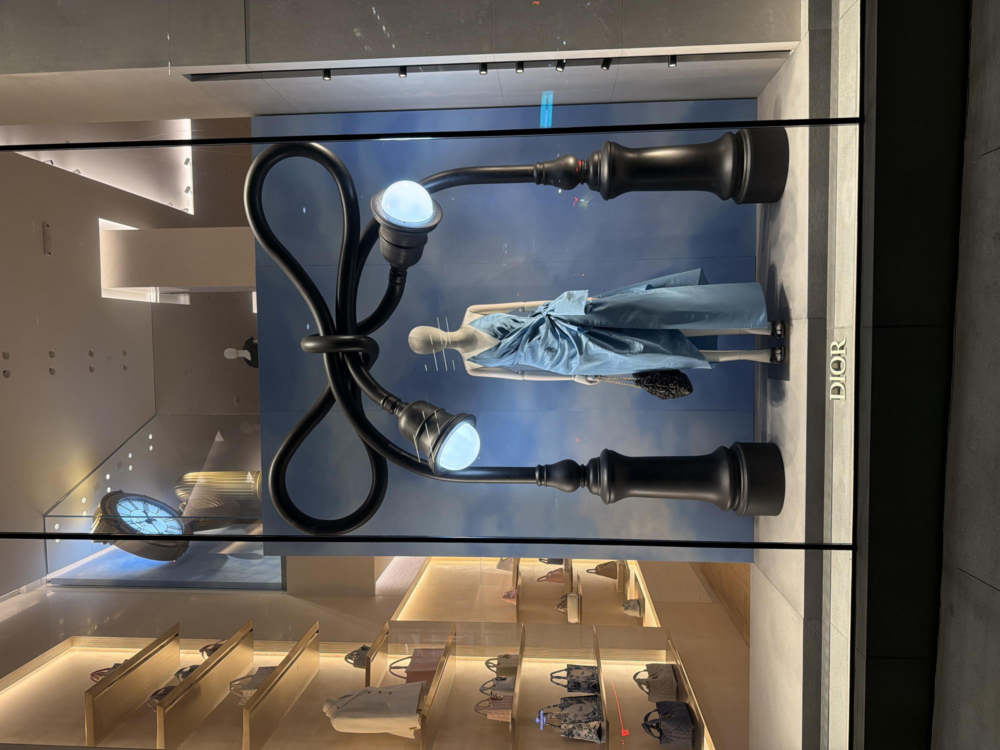 This is a street-level, eye-level, medium shot of a Dior store window display, as seen from the exterior sidewalk of the store at night. The store is located in New York City, USA.
The window display features a mannequin dressed in a light blue, draped dress with a cinched waist and a bow detail. The mannequin is standing in front of a backdrop that appears to be a cloudy sky. Two large, black, sculptural lampposts with glowing white lights frame the mannequin. The lampposts are designed with a loop shape at the top, resembling a ribbon or bow.
To the left of the display, shelves are visible, stocked with various handbags. One shelf displays a white blazer on a form. The shelves and interior lighting create a warm, ambient glow.
A large, ornate clock with a Roman numeral face is visible in the upper background, reflecting in the window. The floor of the display area is a light gray, and the Dior logo is subtly displayed in metallic letters on a dark gray base at the bottom of the window. The overall impression is one of sophisticated luxury and artistic presentation, typical of high-end retail window displays.