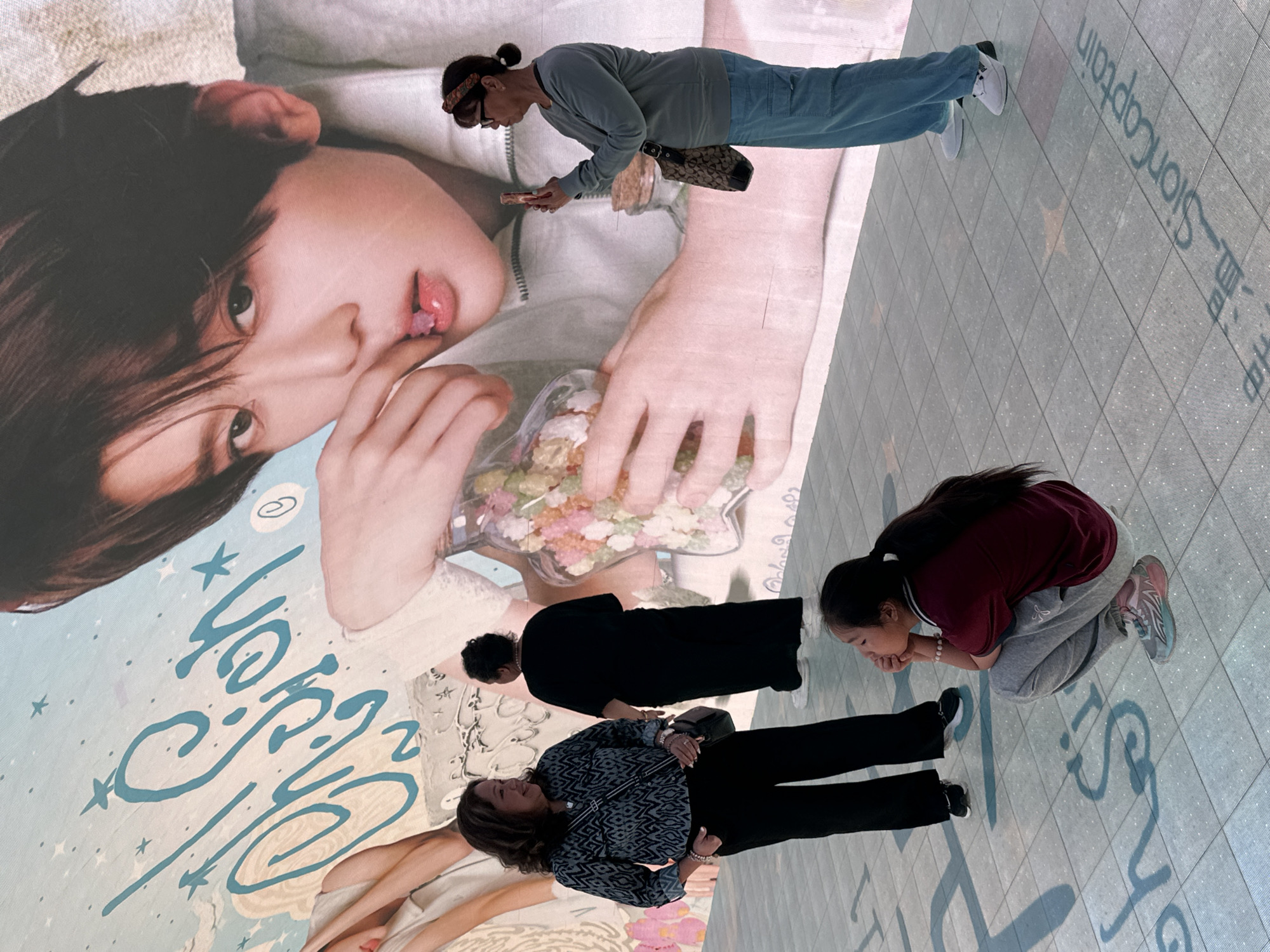 Several people are posing on a large promotional floor advertisement of a young man. The scene is in a building in Yuzhong District, China. One person, an older woman, is standing and holding a phone; the others are crouching and kneeling.