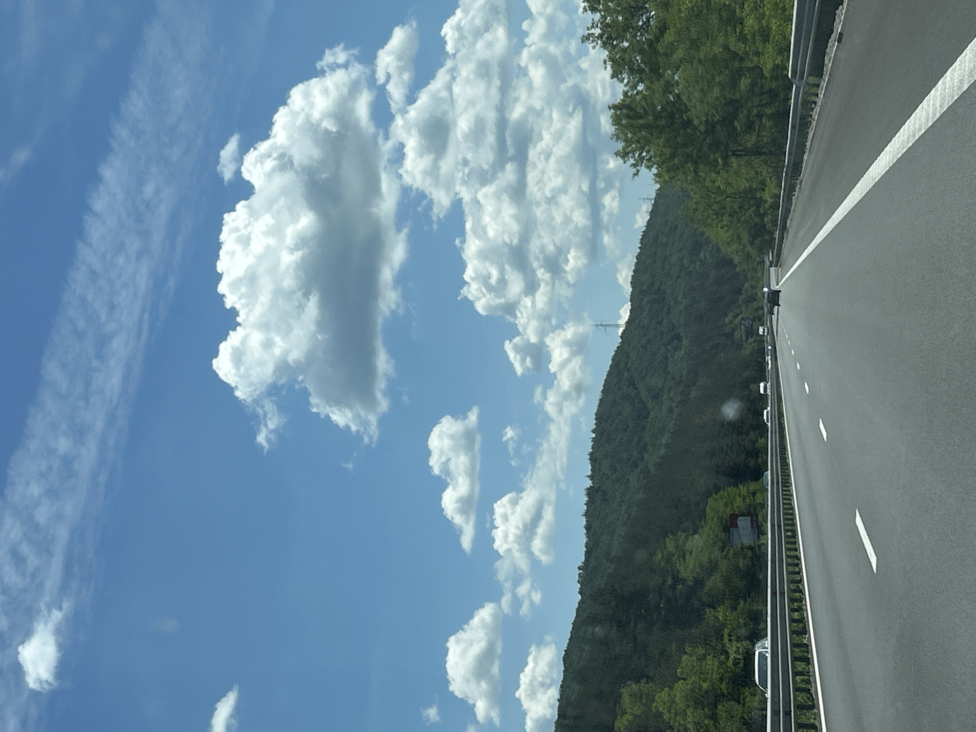 The image depicts a bright, sunny day with a clear blue sky dotted with fluffy white clouds. A highway stretches into the distance, bordered by a metal guardrail on both sides. Lush green trees and a forested hillside dominate the background. Several vehicles, including cars and a white van, are visible on the highway. The scene suggests a typical view from a car driving on a rural highway. The specific location in the city of Rajana, Bulgaria, is not identifiable from the visual information provided.