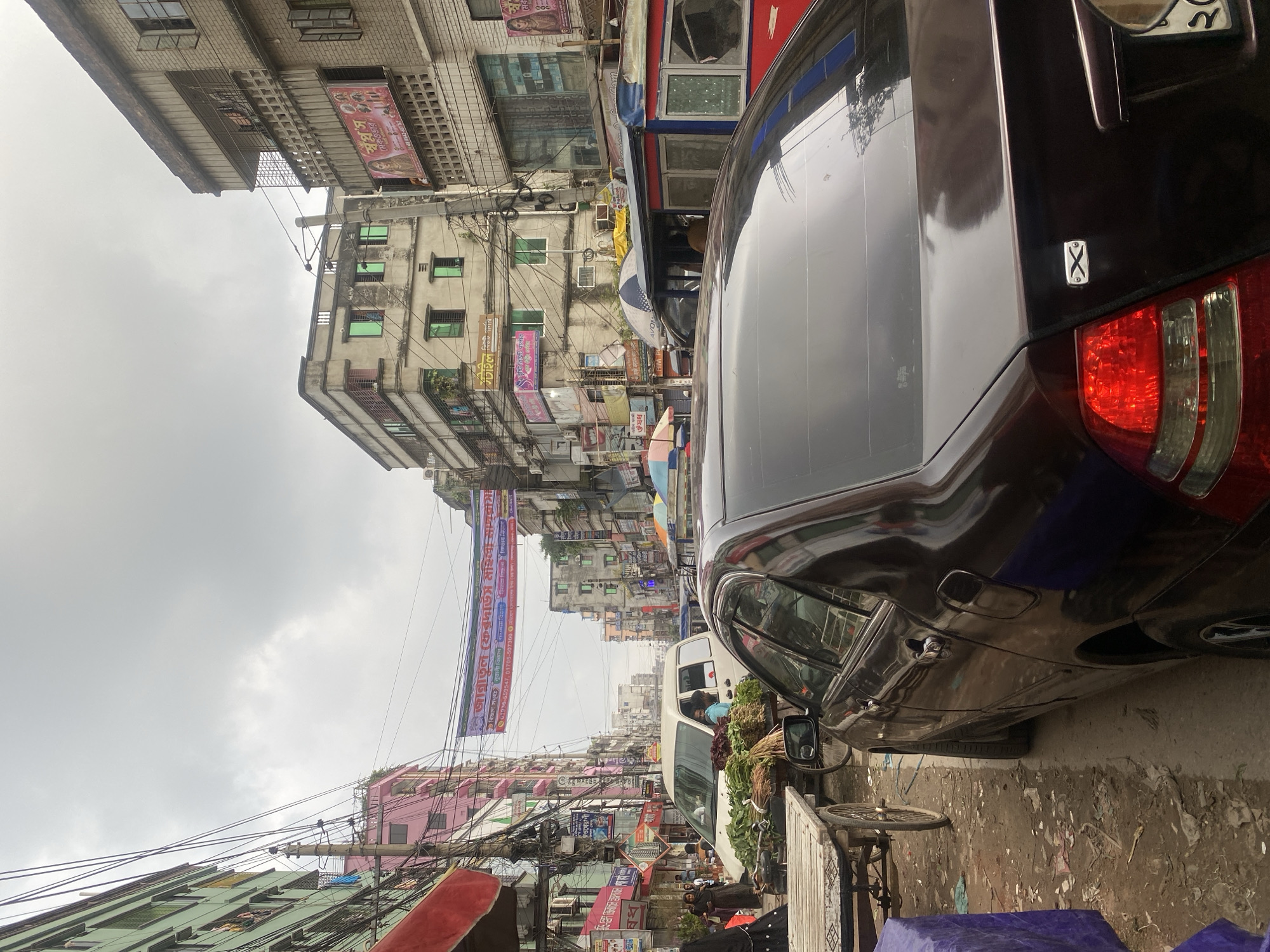 The image depicts a crowded street scene in Dhaka, Bangladesh, characterized by a mixture of vehicular and pedestrian traffic, commercial buildings, and an overcast sky. In the foreground, a dark-colored sedan is parked on the right, with its rear visible, showing a taillight and a license plate. To the left of the sedan, a wooden cart laden with green produce, possibly vegetables or herbs, is positioned on a dirt and debris-strewn roadway.
Behind the cart and sedan, a white van is visible, partially obscuring a person in the driver's seat. Further down the street, a variety of multi-story buildings line both sides, displaying a mix of architectural styles and colors, including pastel pink, green, and grey. Signs in Bengali script are affixed to the building facades, indicating commercial establishments. Power lines and electrical wires crisscross overhead, a common sight in urban Bangladesh.
A banner with text in Bengali is strung across the street between buildings, suggesting a local event or advertisement. In the background, more buildings and a hazy sky complete the urban landscape. Pedestrians are visible walking on the sidewalks and in the street, some of whom are obscured by vehicles. The overall impression is one of a bustling and somewhat congested urban environment typical of Dhaka.