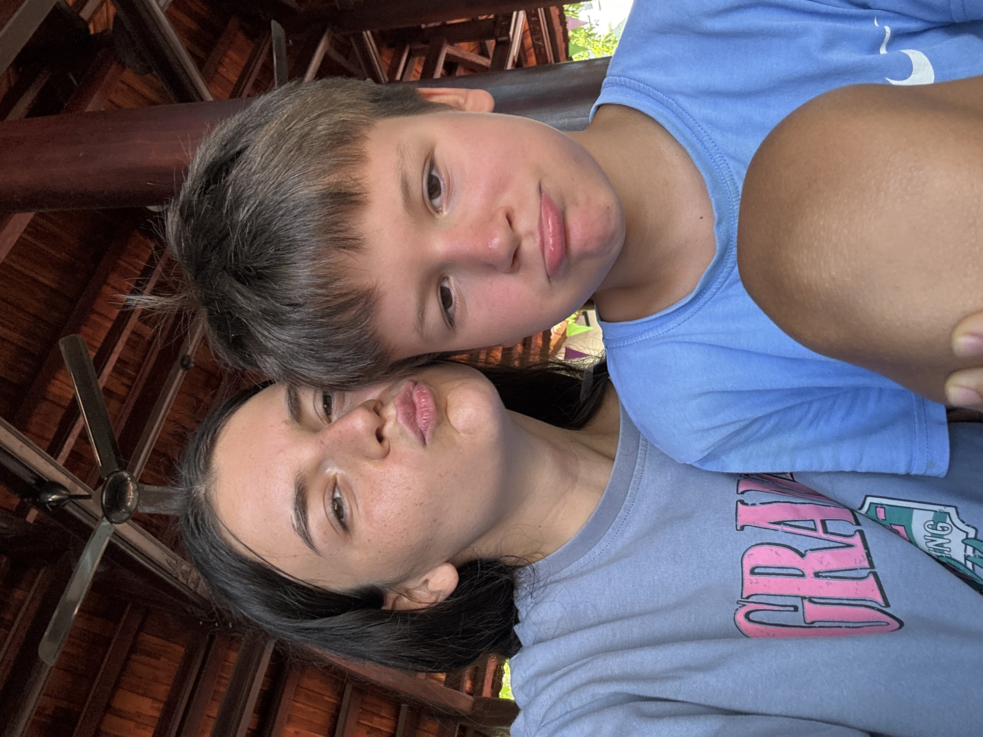 Two individuals are captured in a close selfie under a wooden structure. A fair-skinned boy with short brown hair wears a light blue t-shirt and appears above the other individual. The second individual, a woman with long dark hair, wears a gray t-shirt. The background shows a wooden ceiling and part of a ceiling fan. Given the architectural context, the photo was likely taken in Mũi Né, Vietnam.