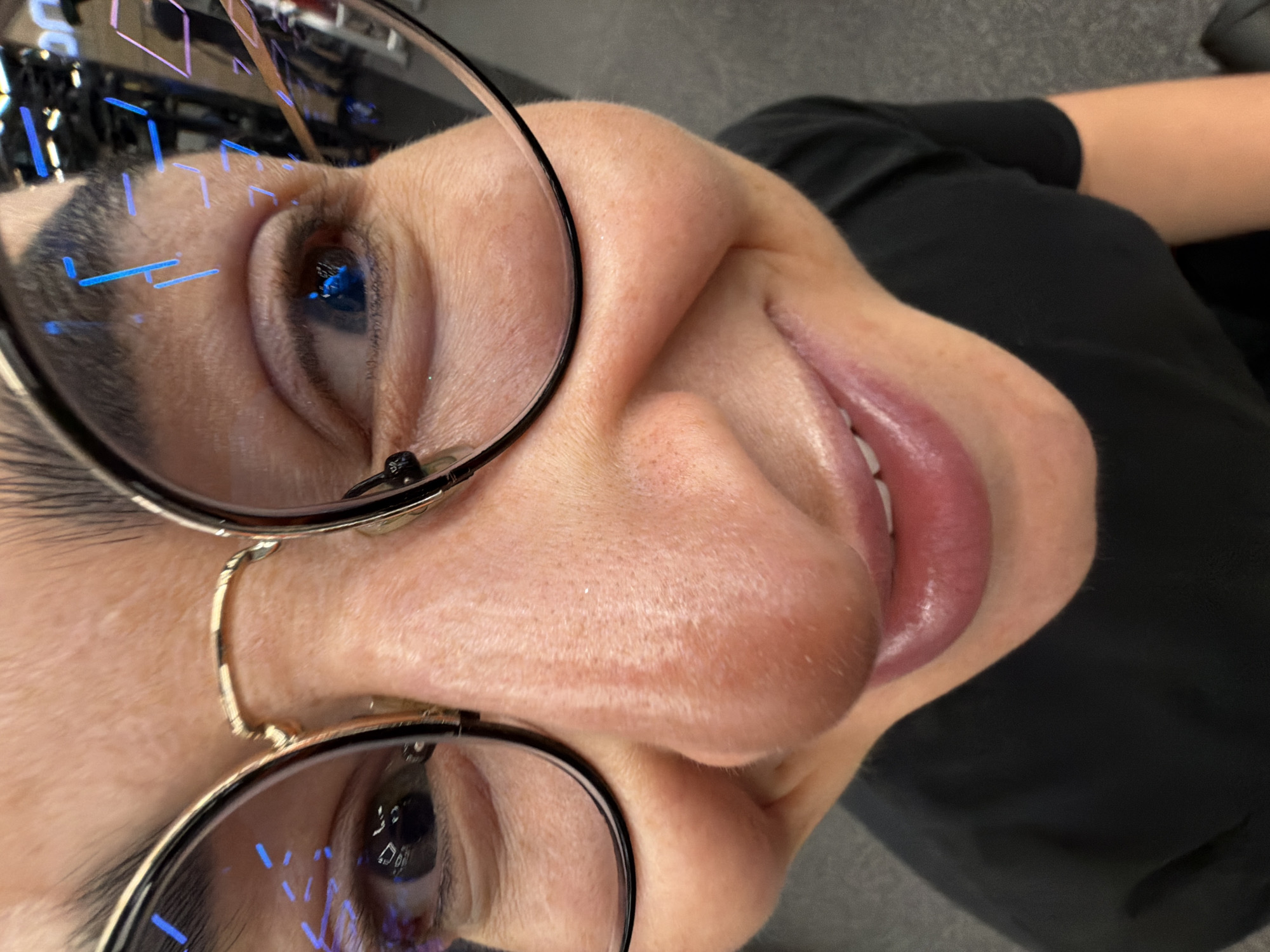 A close-up, slightly distorted, selfie of a person wearing eyeglasses. The person is fair-skinned with dark hair and is wearing a black top. Reflections of blue and white light are visible in the lenses of the eyeglasses. The background is out of focus and appears to be a gray surface. There is no indication of this image being in Saint Petersburg, Russia.