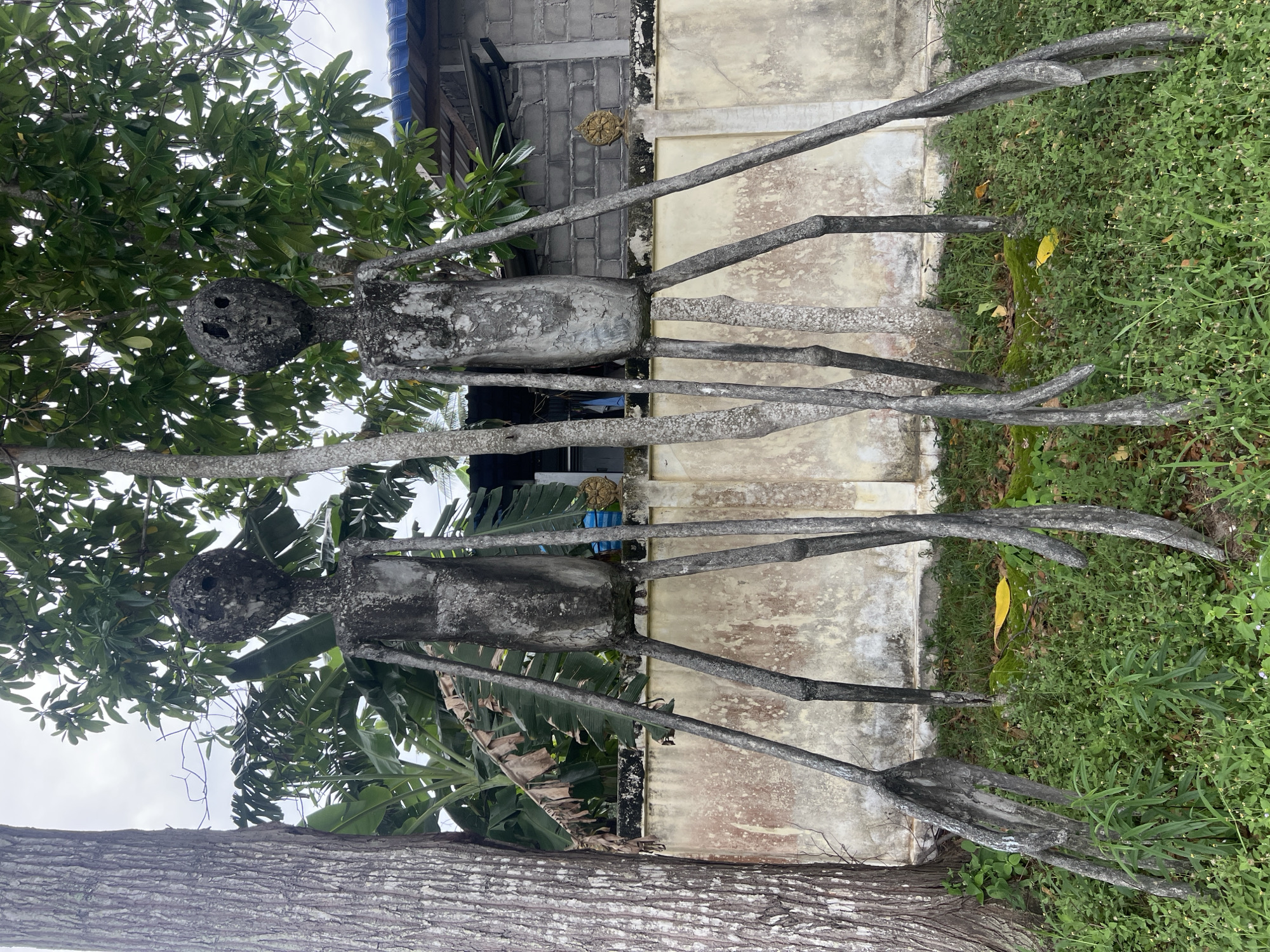 Two abstract, humanoid concrete sculptures stand in a grassy area in front of a weathered, cream-colored wall. The sculptures are grey and have a rough, textured surface. Each sculpture has a tall, slender torso and extremely long, thin legs that splay outwards. Their heads are oval-shaped with two dark, circular openings for eyes and a simple slit for a mouth. The sculpture on the left is slightly obscured by a thick tree trunk. Behind the wall, there is lush green foliage, including banana leaves, and a building with a blue roof. The sky is overcast. The scene appears to be outdoors in a tropical climate, likely within the Ko Pha-ngan District of Thailand.