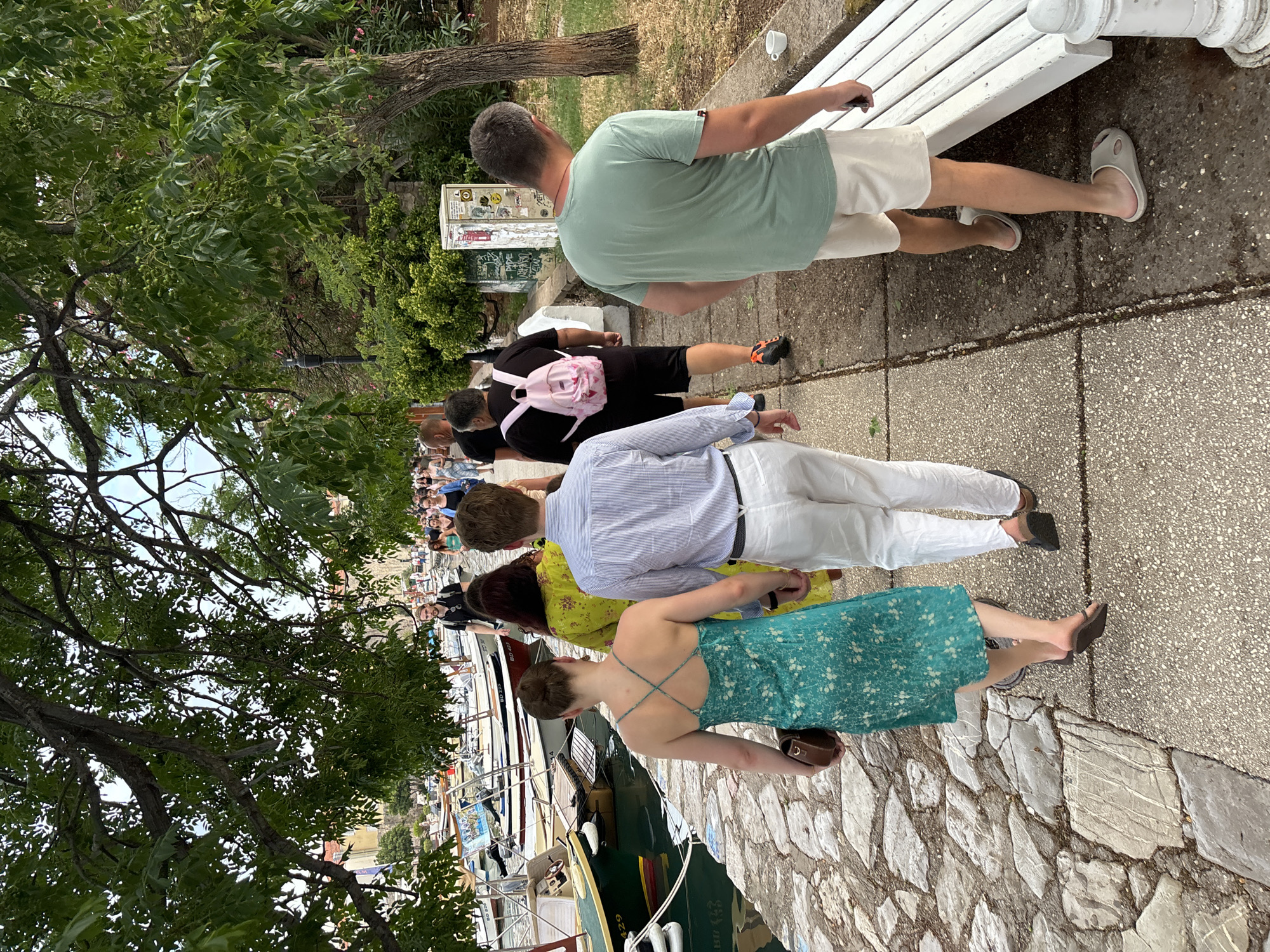 In Budva, Montenegro, a group of people are walking along a waterfront promenade. The scene is outdoors, with trees and a stone walkway visible. Several boats are docked along the water's edge.
The individuals are casually dressed, suggesting a relaxed atmosphere. One person is wearing a light green t-shirt and white shorts, while another is in a black t-shirt and shorts, carrying a pink backpack. Further down the line, a person in a blue and white striped shirt and white pants is walking. A woman in a yellow dress and another in a turquoise floral dress are also present. Their body language suggests they are moving in the same direction, perhaps as a group or part of a larger flow of people.
The walkway is paved with stone, and a white bench is situated along the side. The overall impression is a pleasant day, likely during daylight hours, with moderate weather conducive to outdoor activities. The presence of boats indicates a coastal or harbor setting. There is no visible text that provides specific information.