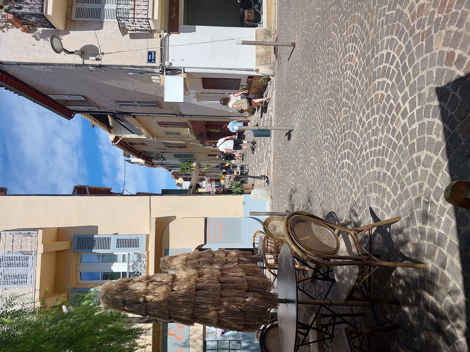 The image depicts a sunlit street scene in Antibes, France, likely in the Old Town due to the cobblestone pavement and narrow street flanked by buildings. In the foreground, outdoor seating for a cafe or restaurant is visible, with wicker chairs and a round table partially obscured by a large, straw-like umbrella. The buildings lining the street have traditional architecture, featuring light-colored facades, shuttered windows, and balconies. A street sign reading "RUE du FERSEN" is clearly visible on the right side of the image. Several people are walking along the street, some in casual attire, suggesting a pleasant, daytime atmosphere. The overall impression is one of a typical European street scene, with a focus on pedestrian activity and outdoor dining spaces.