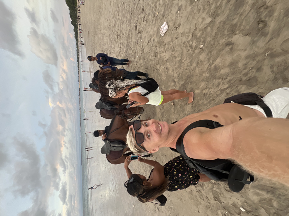The scene depicts a sandy beach in Jimbaran, Indonesia, under an overcast sky during daylight hours. In the immediate foreground, a shirtless Caucasian male with blond hair and sunglasses positioned on his head is taking a selfie, smiling directly at the camera. He wears white shorts, a black backpack, and a black fanny pack.
To his left, a woman with dark hair tied back, wearing a dark floral strapless top and dark sunglasses on her head, is holding a smartphone in an upward position, seemingly capturing an image or video. A black shoulder bag hangs from her left shoulder.
To the right of the selfie-taker, a second woman with long, light-colored braided hair is holding a smartphone, also appearing to capture media. She wears a light-colored top, shorts, and a black shoulder bag, and is barefoot. A pair of white sandals are visible on the sand to her right.
Behind these individuals, three saddled brown horses are present. Two male attendants are positioned near the horses. One male, wearing a dark t-shirt, is adjusting a saddle on the leftmost horse. A second male, wearing a dark blue long-sleeved shirt and a baseball cap, stands near the two rightmost horses, holding a dark object.
The background reveals a wide expanse of beach meeting the ocean. Numerous individuals are scattered along the shoreline and in the shallow water, engaged in various activities such as walking or wading. Distant land features and structures are faintly visible along the horizon. The sky is largely covered with light and dark clouds.