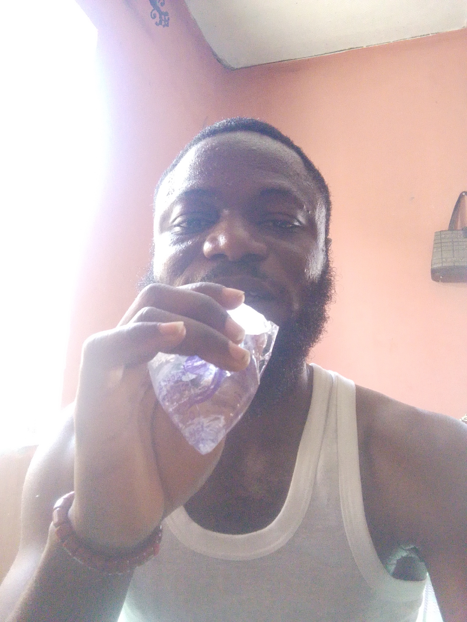 A man with dark skin, a beard, and wearing a white tank top holds a small, transparent plastic packet of liquid to his mouth. He is looking at the camera. He is wearing a red bracelet on his left wrist. Behind him is a light orange or peach-colored wall. In the upper right corner of the frame, a handbag is hanging on the wall. Based on the request, this image may be taken in the city of Lokoja, Nigeria.