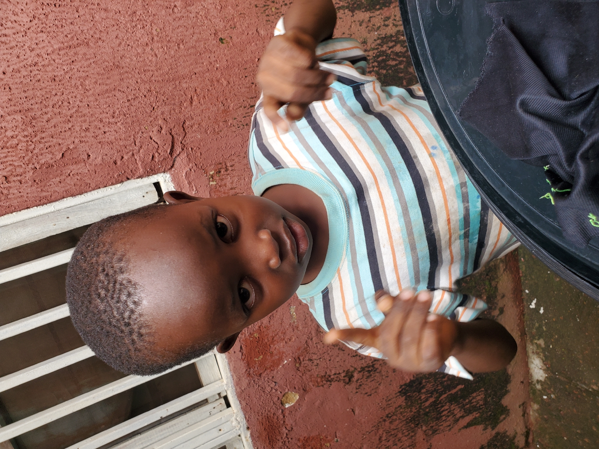 A young Black child, likely a boy, stands in front of a textured, reddish-brown wall. The child is wearing a striped t-shirt with bands of blue, white, orange, and black. His right hand is raised, index finger pointing forward, as if making a demand or accusation. His left hand is also raised, with the index finger pointing. The child's gaze is directed slightly off-camera. To the left of the child, there appears to be a white barred window. In the foreground, a portion of a black, circular object, possibly a bucket or basin, is visible, with a piece of black fabric draped over it. The ground beneath the child and the wall is a rough, uneven concrete surface. The overall lighting suggests an outdoor setting, possibly during the day, with the sun casting some shadows. The scene's context within Jos, Nigeria, is not directly indicated by the visual elements.