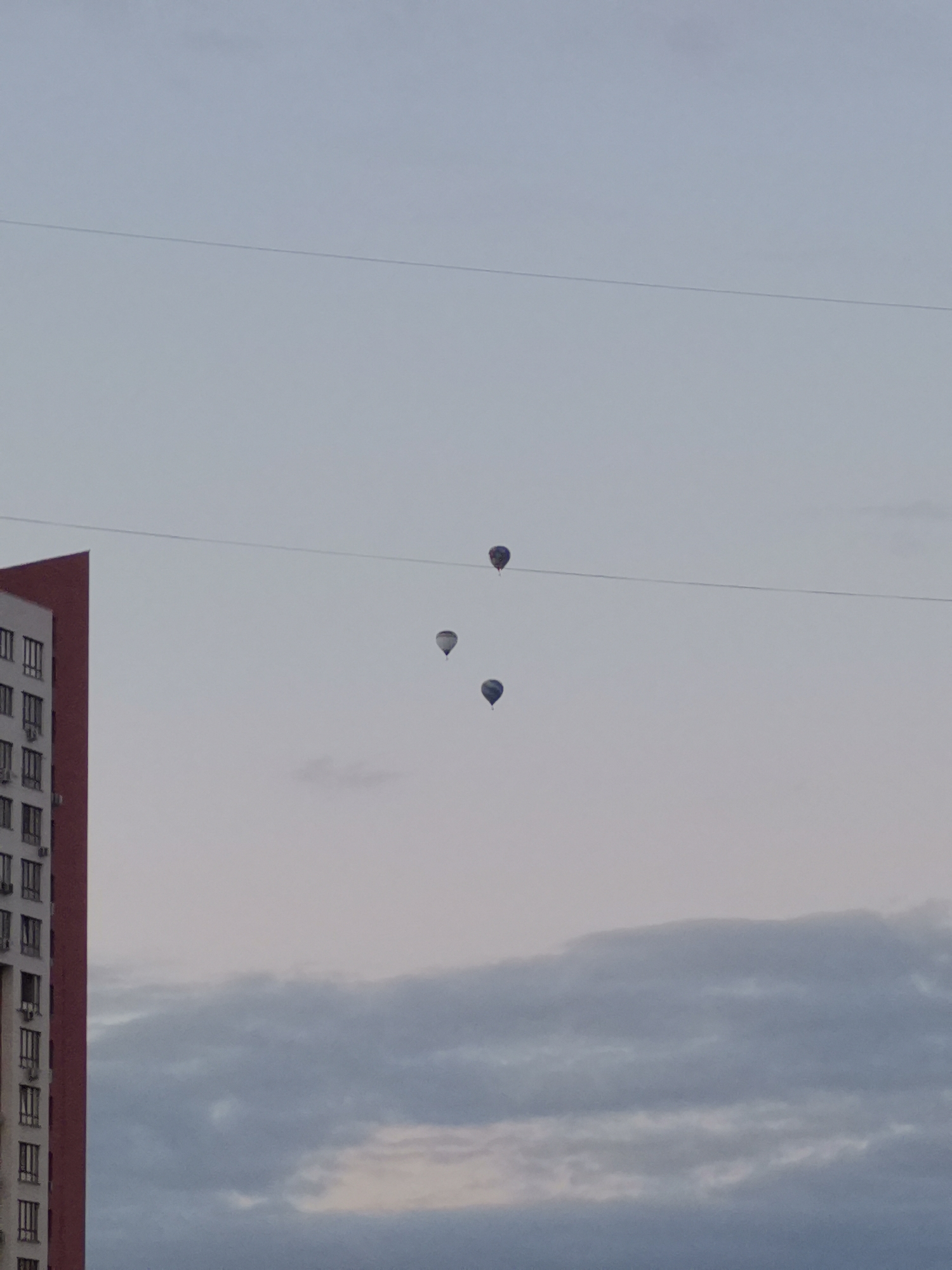 The image captures a view of the sky above Kemerovo, Russia, with a tall, modern building on the left side of the frame. The building features a red facade and numerous windows, indicating a residential or commercial structure. Three hot air balloons are visible in the sky, suspended at different altitudes. They appear to be dark in color and are ascending or drifting. A thin, horizontal wire traverses the sky across the frame, intersecting the path of the balloons. The sky itself is a pale blue, transitioning to a lighter hue towards the horizon, with scattered clouds present in the lower portion of the image. The overall scene suggests an outdoor activity or event taking place in the city.