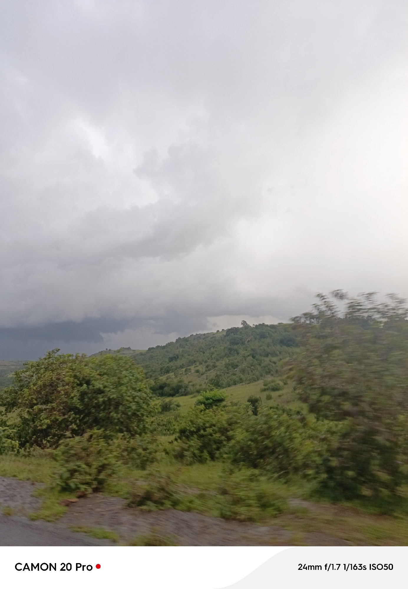 A cloudy sky hangs over a lush, rolling green landscape, likely the city of Nguroje, Nigeria. The foreground is dominated by a blurred image of the terrain, suggesting movement, possibly from a vehicle. Dark, heavy clouds gather on the left side of the sky, hinting at impending weather. The middle ground features a hillside covered in dense green vegetation and scattered trees. The far background is a hazy expanse of sky and distant hills. Text at the bottom of the image reads "CAMON 20 Pro" and "24mm f/1.7 1/163s ISO50." No people or specific objects are clearly identifiable due to the motion blur.