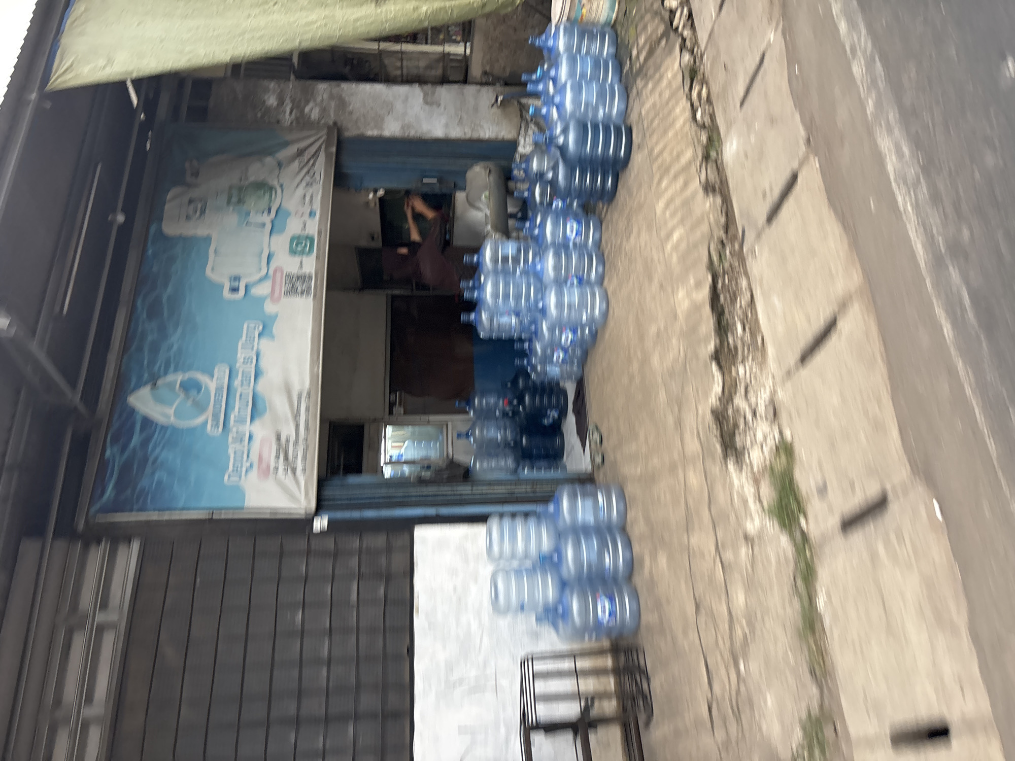 This is a medium shot, street-level photograph taken from a slightly elevated perspective, depicting the storefront of a water refilling station located in Bekasi, Indonesia. The storefront features a blue awning with a banner displaying a water drop logo and images of water bottles. In front of the store, numerous large, clear plastic water jugs are neatly stacked on the concrete ground. Some jugs are on the sidewalk, while others are closer to the street. The building's facade is constructed with dark horizontal siding, and a metal shelf unit is visible to the left of the entrance. Through the open doorway, a person wearing a dark brown headscarf and shirt can be seen behind a counter, engaged in activity. A green tarp hangs from the right side of the awning. The street in front of the store has a concrete curb and drainage system. The overall scene suggests a typical commercial establishment in an urban Indonesian setting.