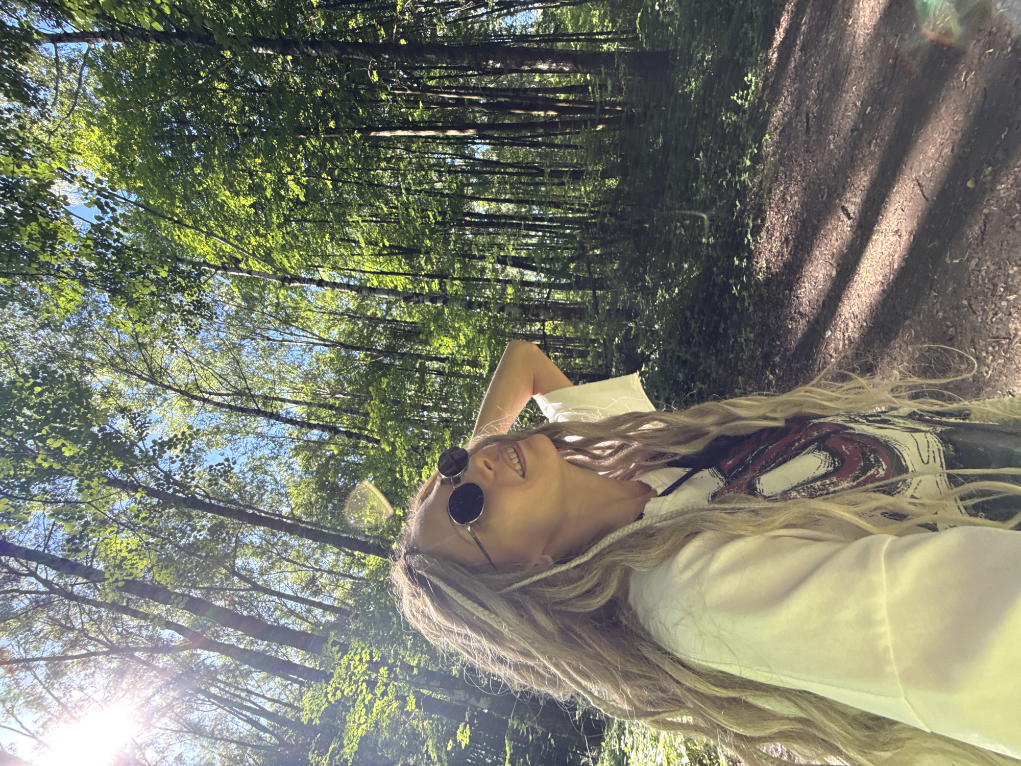 The image depicts a female individual in a sunlit forest in Rusanovo, Russia. The woman, positioned on the left side of the frame, has blonde-to-light-brown hair styled with a mix of dreadlocks and wavy textures. She wears round, dark-lensed sunglasses and a light-colored, long-sleeved shirt with a partially visible graphic print on the front. Her head is tilted upwards and to her left, with a smile on her face, as she looks towards the sky through the tree canopy. Her right hand is raised, resting on the back of her head or neck. The background consists of numerous tall, slender trees with lush green foliage, indicating a dense wooded environment. The sky is visible as clear blue through gaps in the leaves. Strong sunlight enters from the upper left, creating distinct lens flares and bright highlights on the leaves. The ground is covered with dirt and sparse undergrowth, and a shaded dirt path is visible on the right side of the image, marked by elongated shadows from the trees.